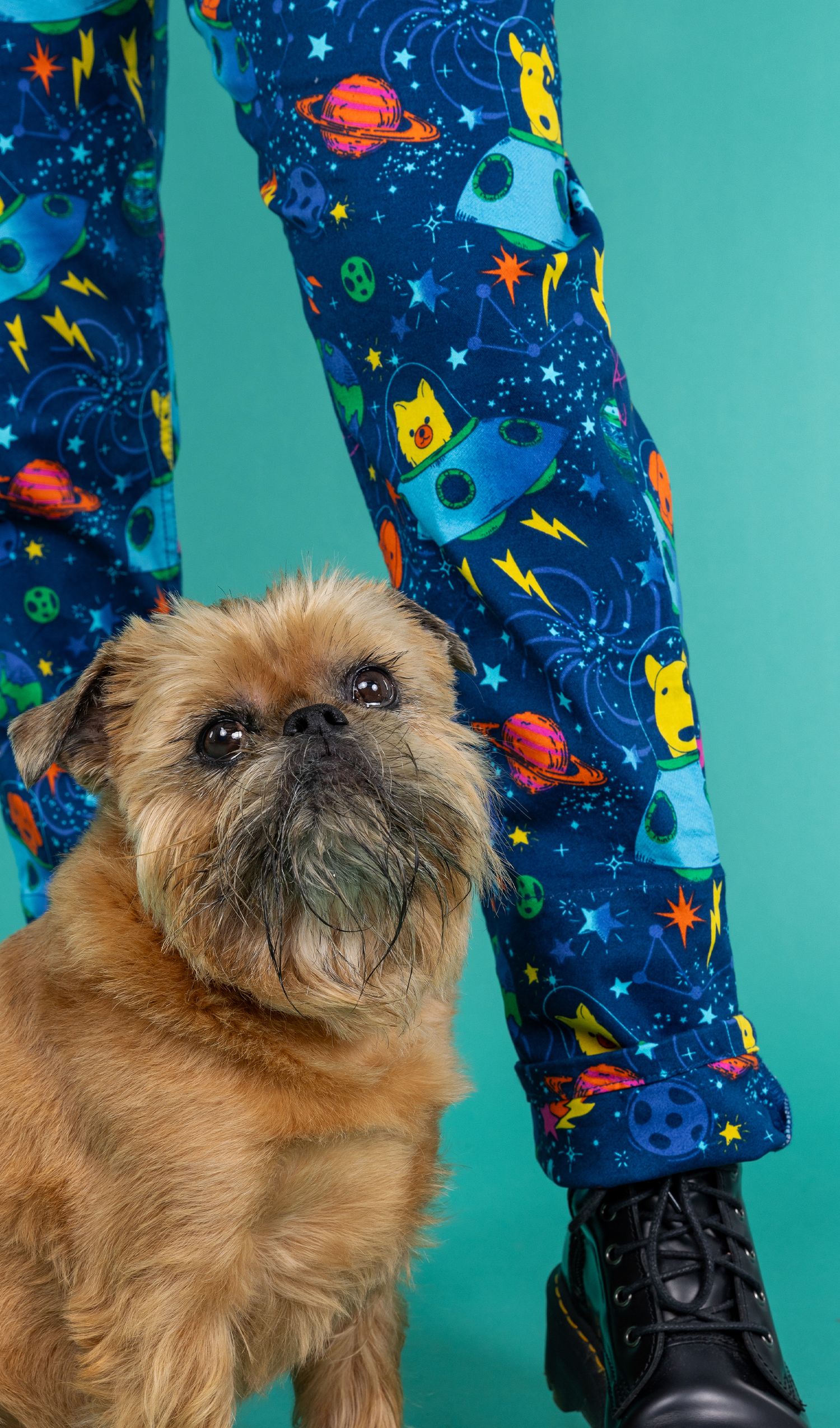 Brian a Brussels griffon dog is sat by the feet of someone wearing dungarees with a space print he is looking into camer