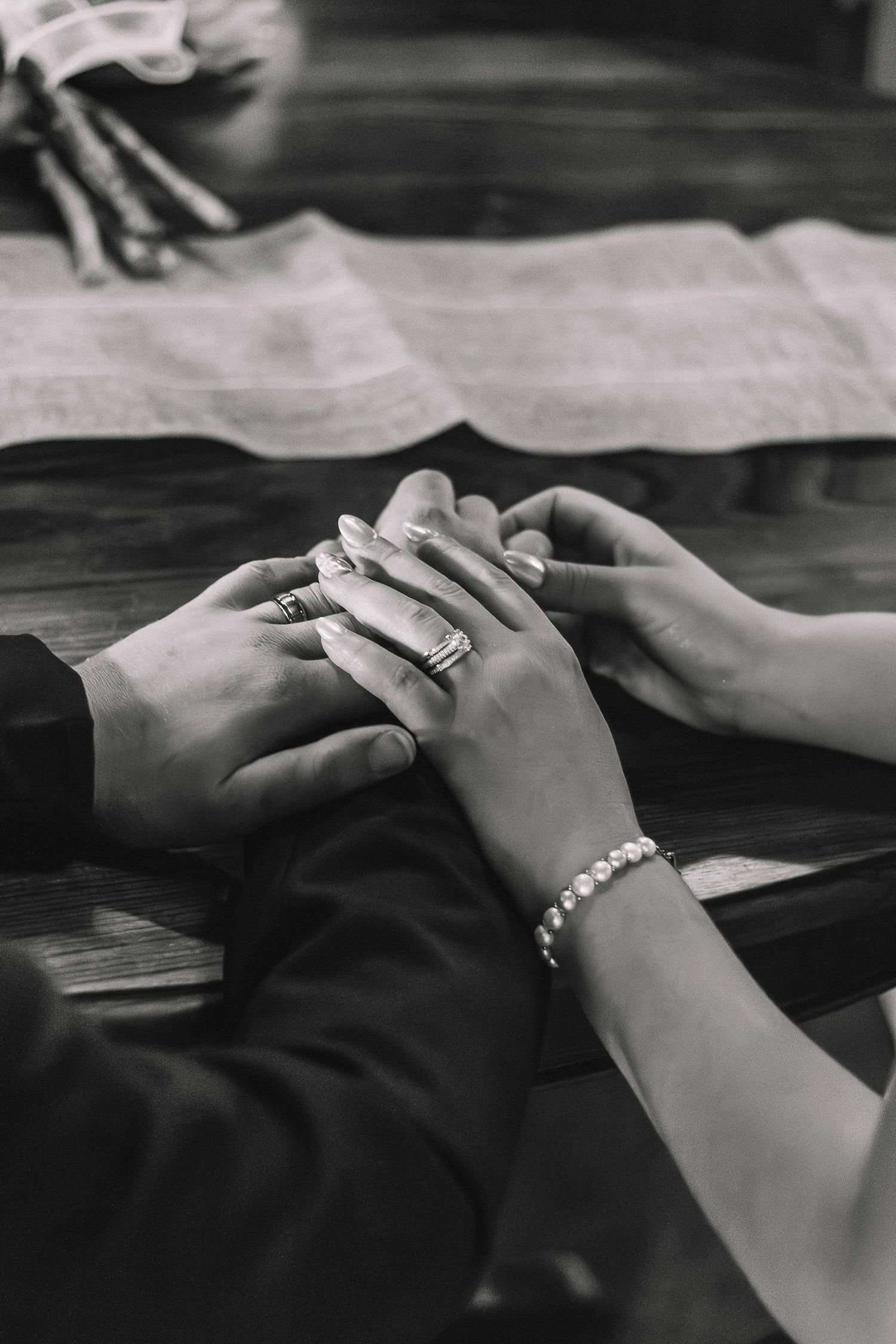 Mood-lit black and white detail photo of a bride and groom holding hands at their wedding table.