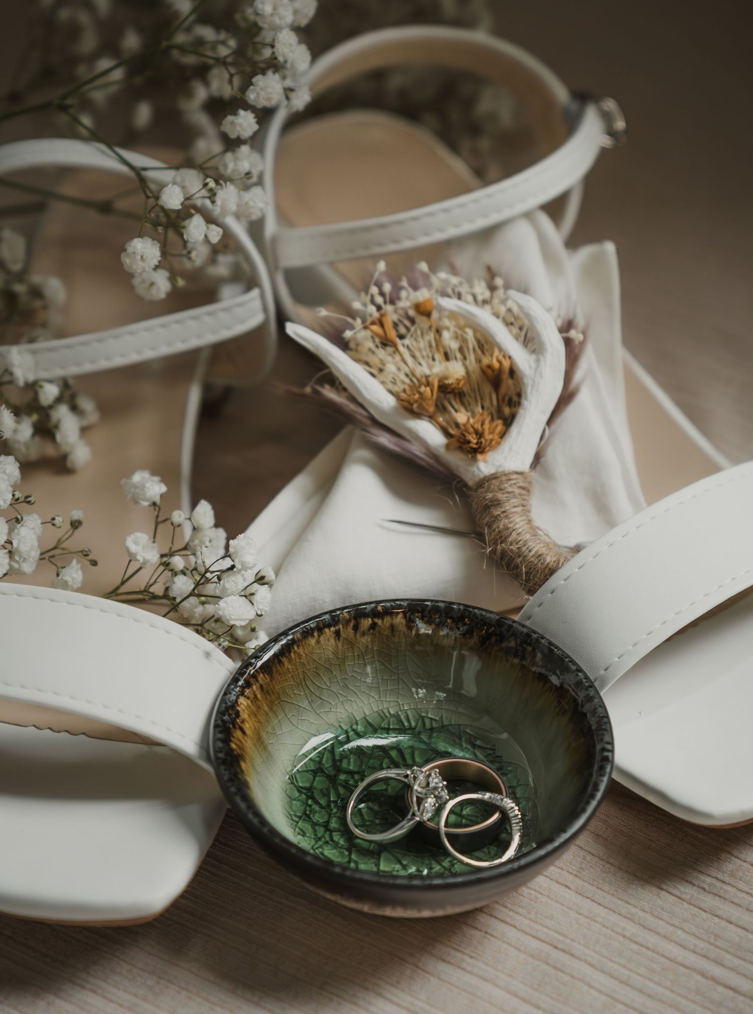 Wedding rings displayed in decorative green dish with white flowers and bridal shoes.