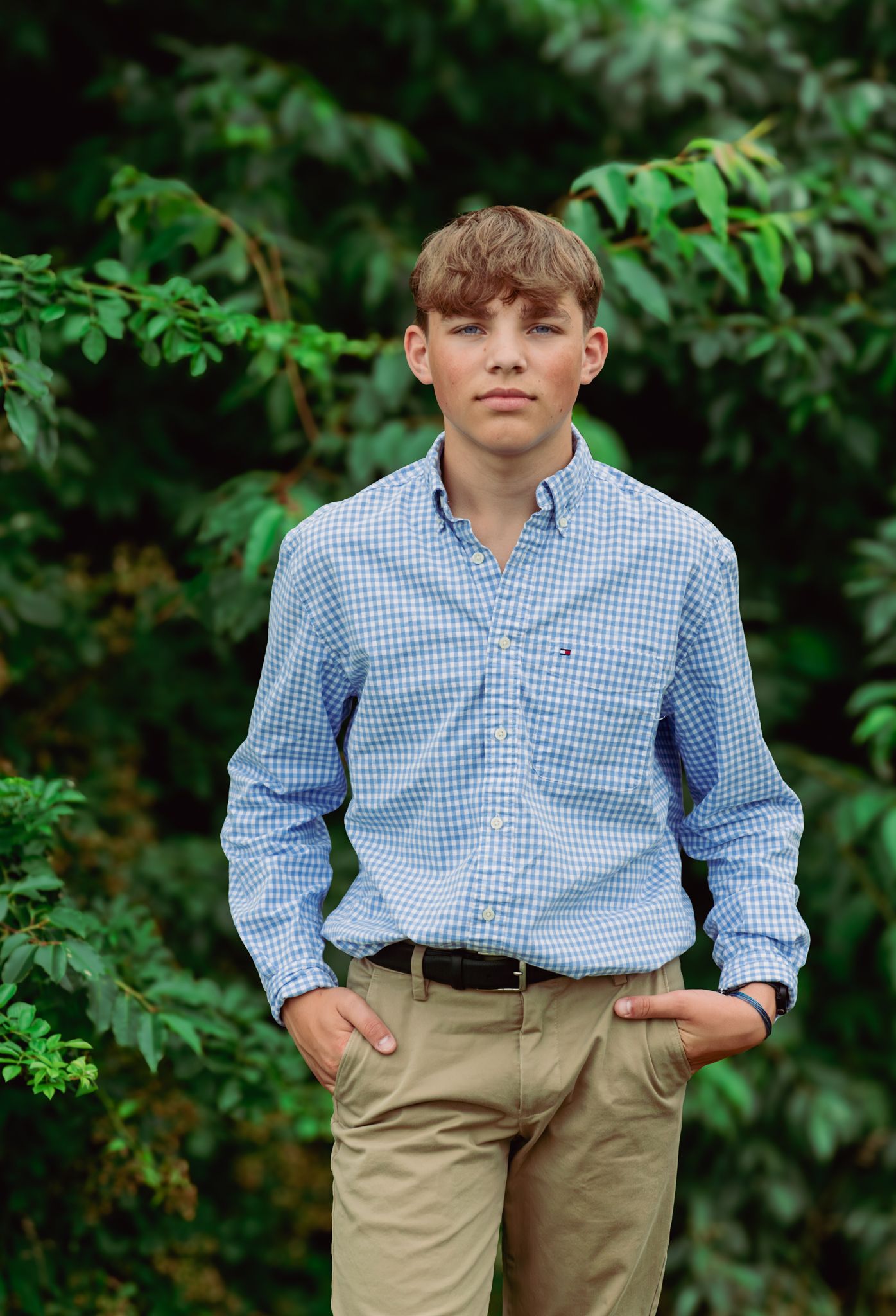 Young person in checkered blue shirt and khaki pants poses against green foliage.