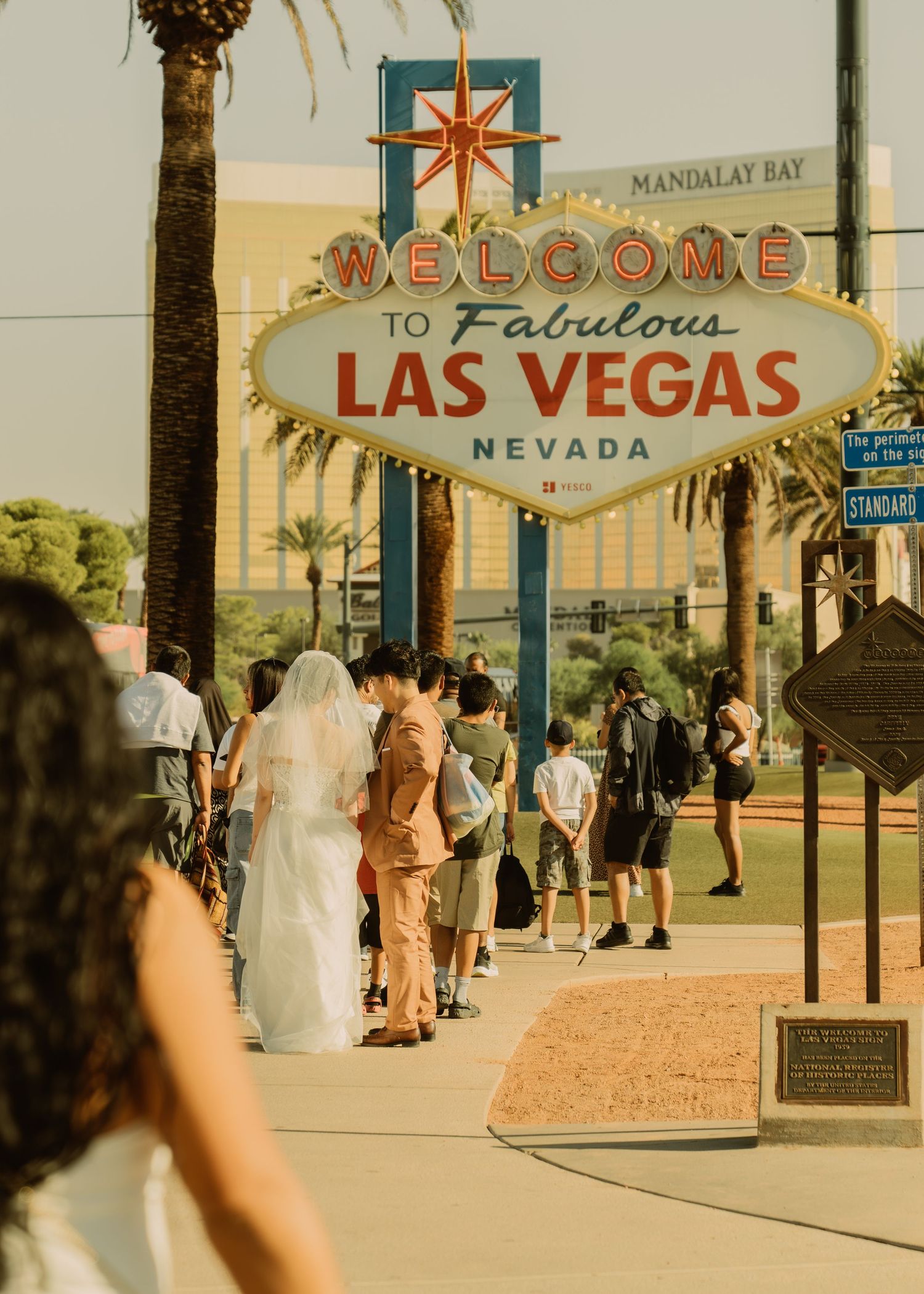 Iconic Welcome to Las Vegas sign with palm trees and visitors gathered beneath it.