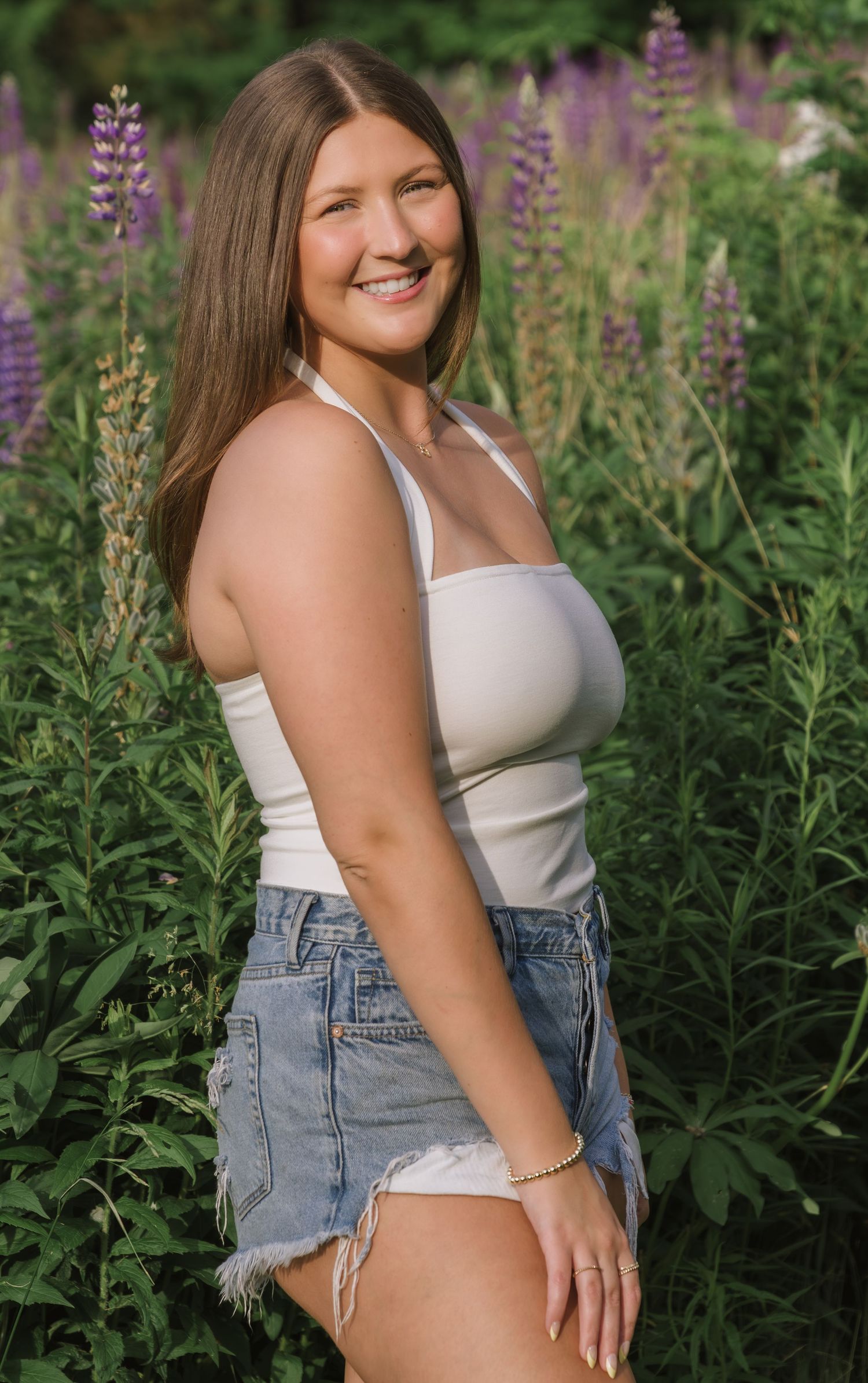 A person in a white tank top and denim shorts poses outdoors among blooming purple lupine flowers in a garden setting.