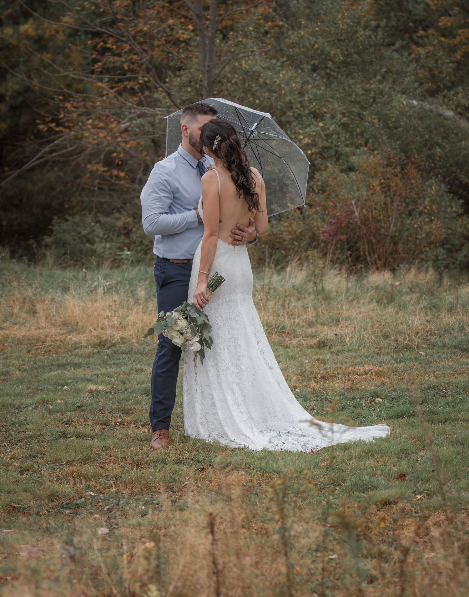 Couple poses in autumn field with long white wedding dress trailing behind.