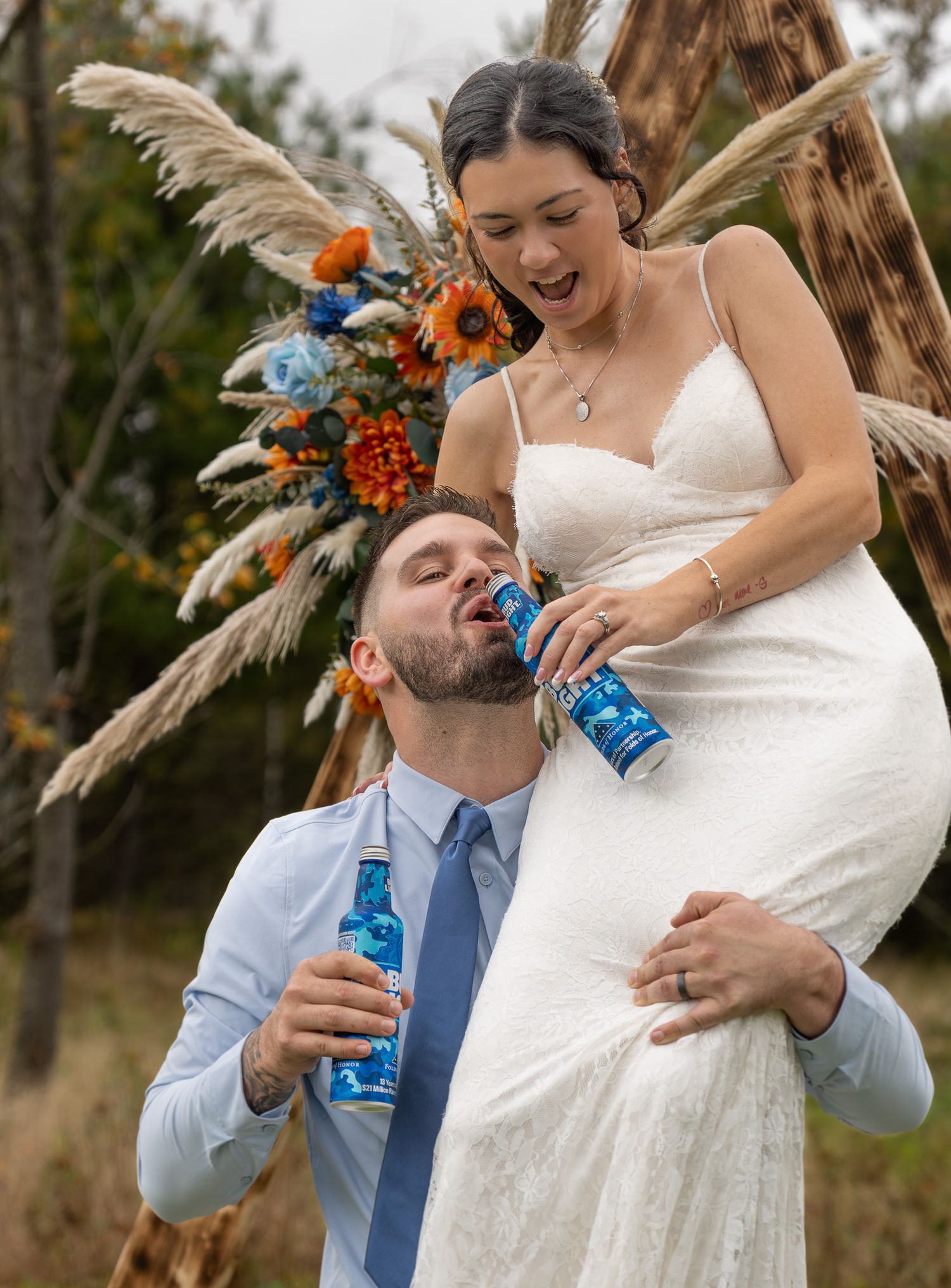 Newlyweds sharing playful drink moment at rustic outdoor wedding setting.