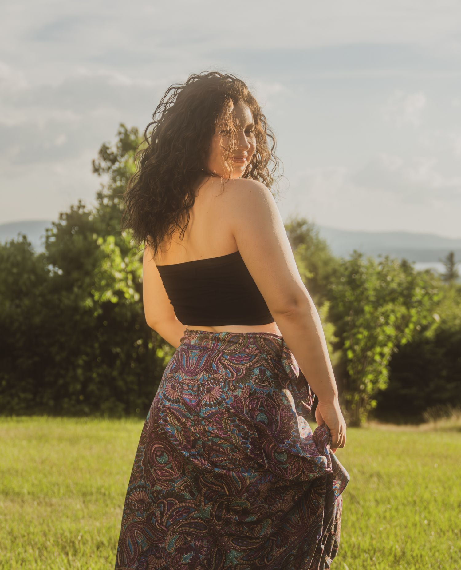 Person wearing a floral print dress twirls gracefully in a sunlit meadow with green trees in the background.