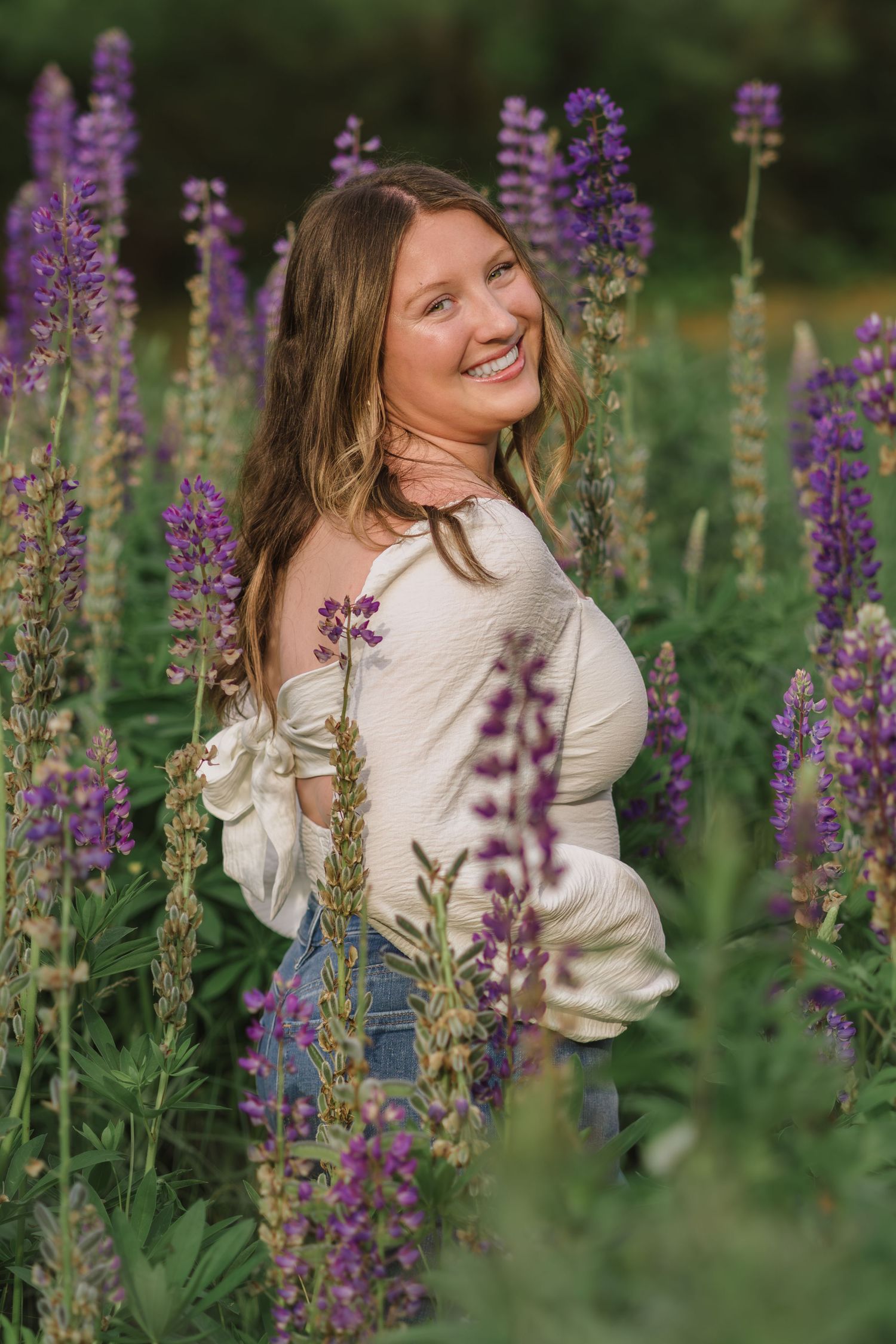A person in a white sweater poses joyfully among blooming purple lupine flowers in a summer garden.