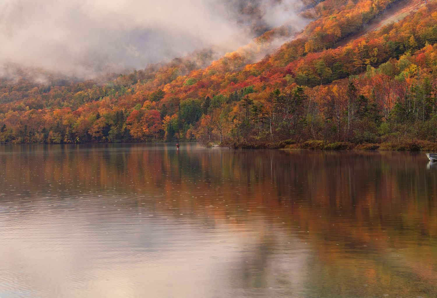Panoramic view of a lake surrounded by autumn trees with vibrant orange and red foliage reflecting in calm waters.