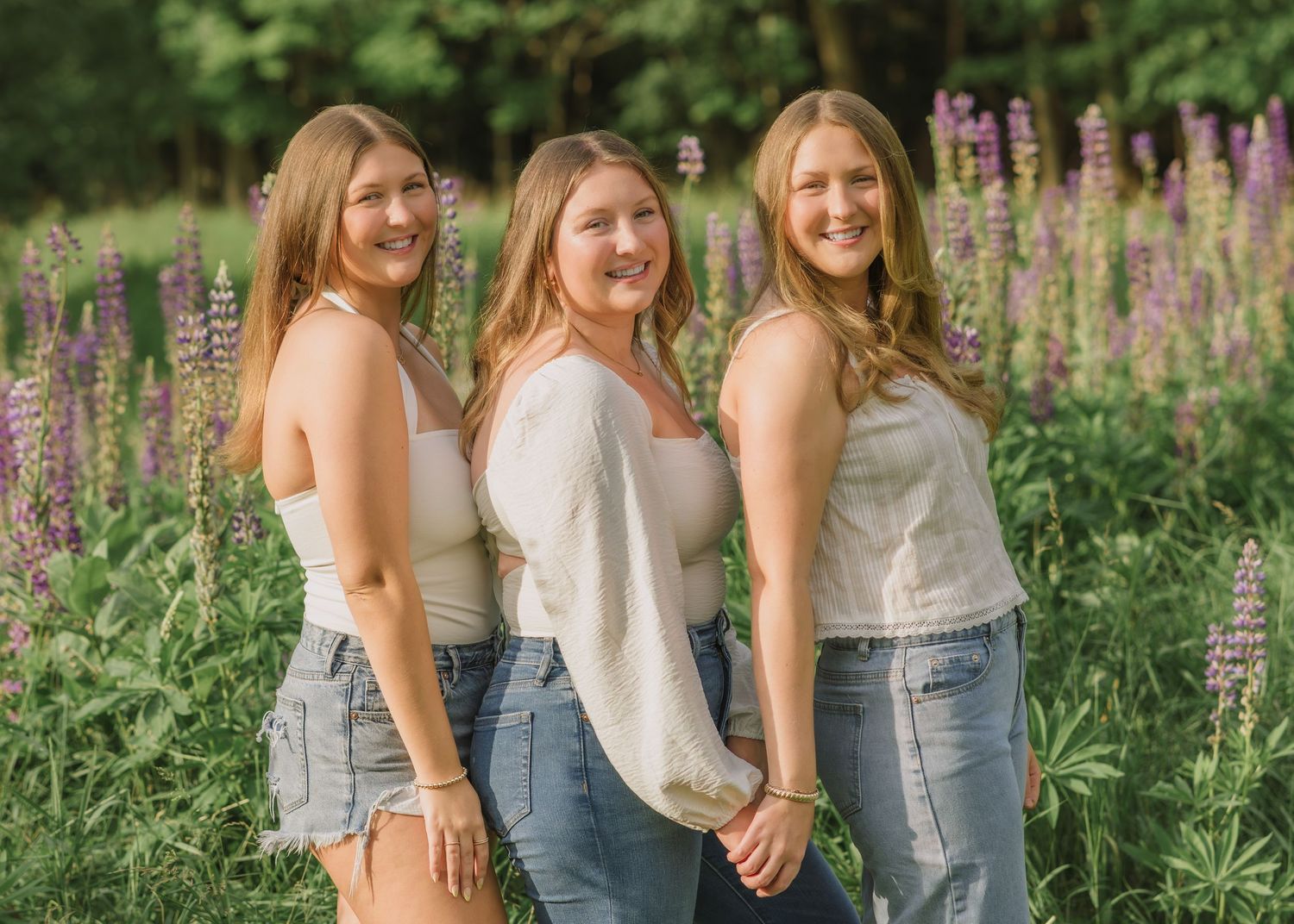 Three women in casual attire pose together in a garden of purple lupine flowers.