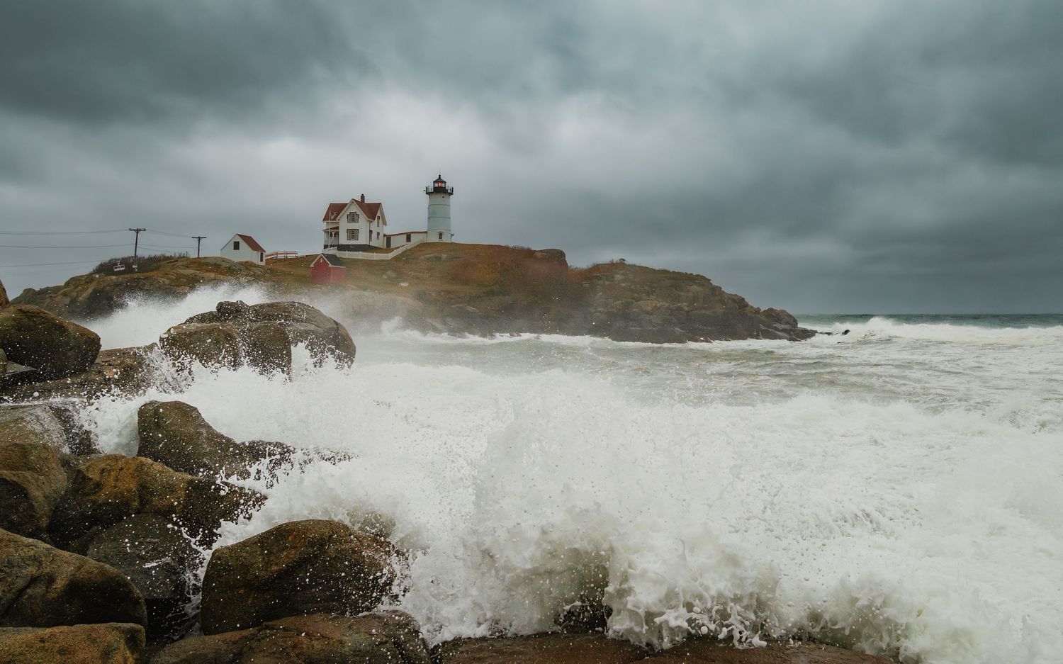 Dramatic waves crash against rocky cliffs beneath stormy skies at a coastal lighthouse.