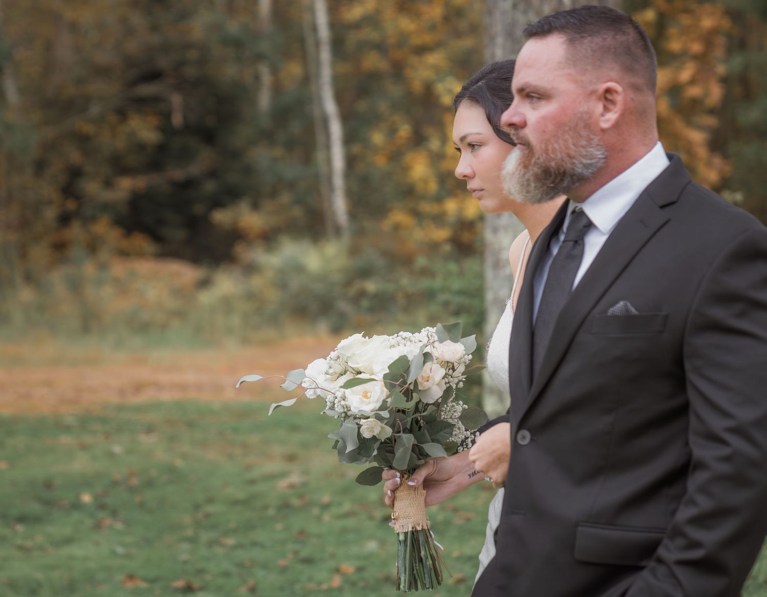 Wedding party stands together outdoors during autumn with bouquets against a backdrop of fall foliage.