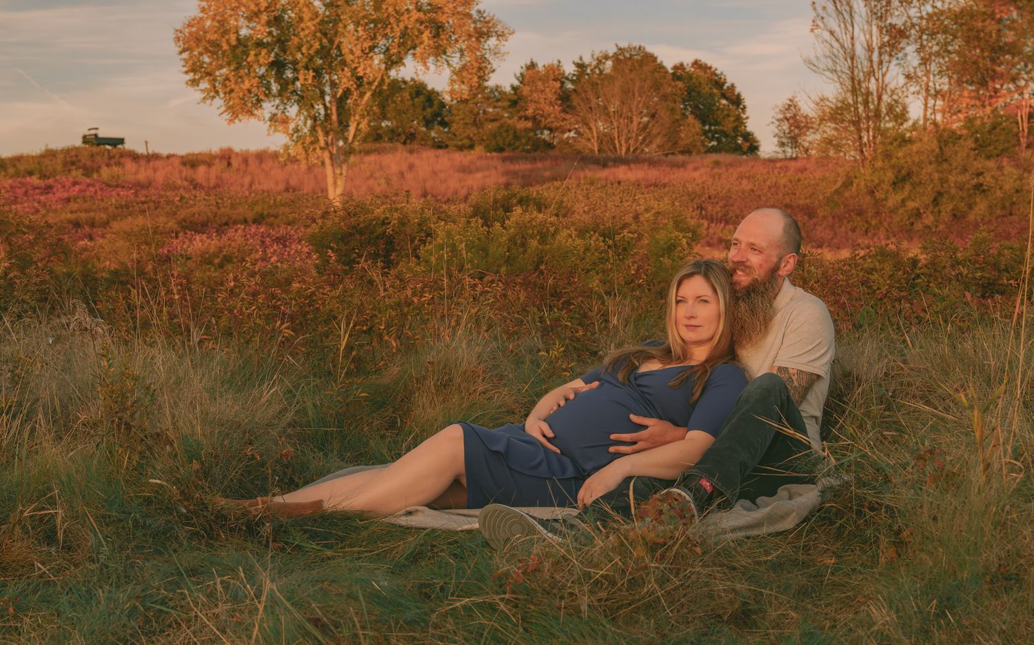 A couple lounges in a field during golden hour with warm autumn colors surrounding them.