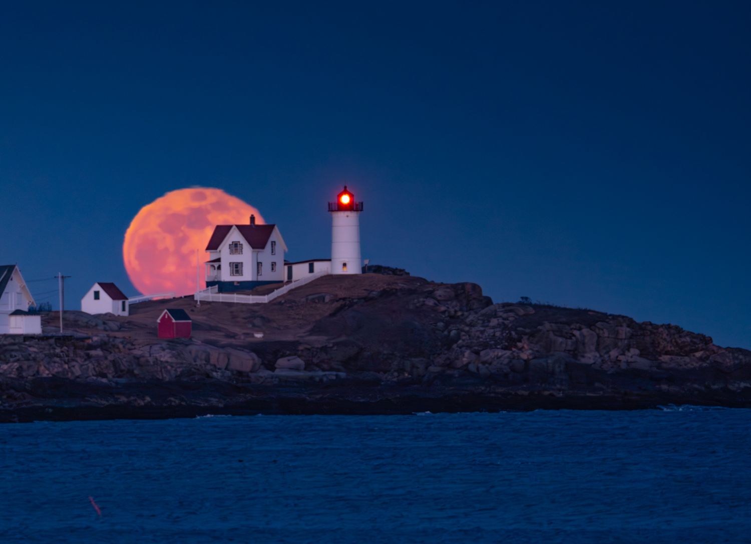 Full moon rising behind Nubble Lighthouse in Maine during blue hour.
