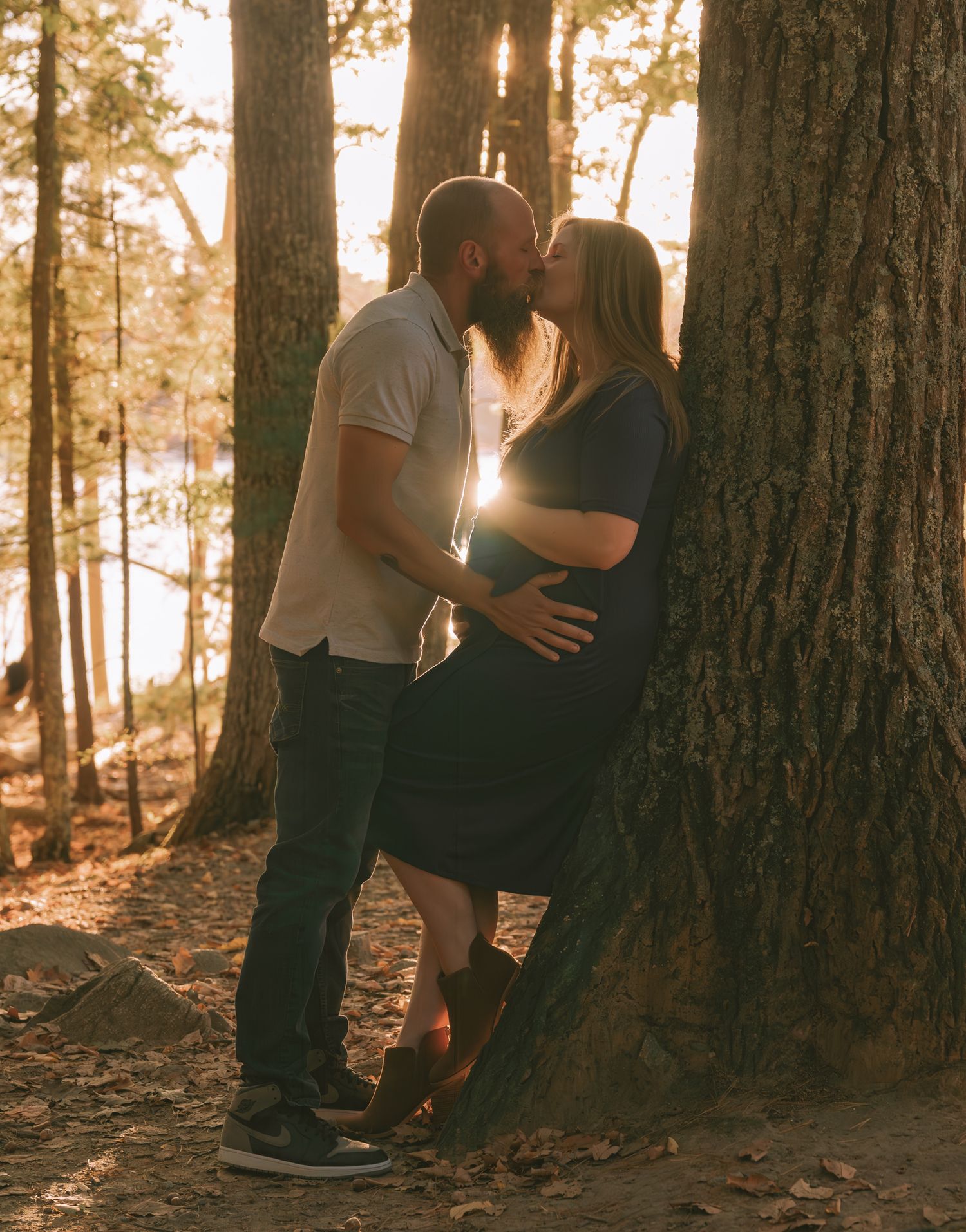 Intimate couple's portrait against tree trunk with sunlight streaming through forest.