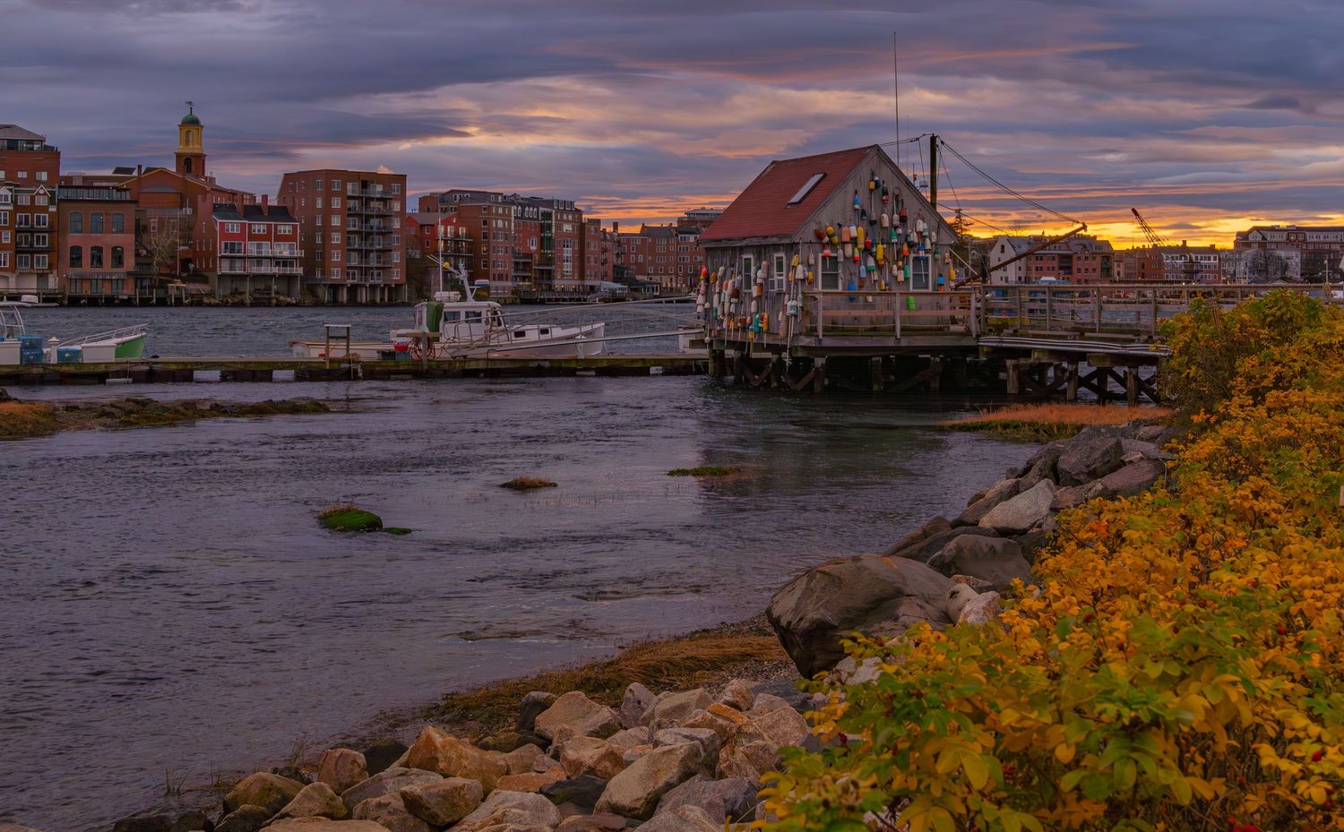Waterfront cityscape with autumn foliage and dramatic sunset clouds reflecting on harbor.