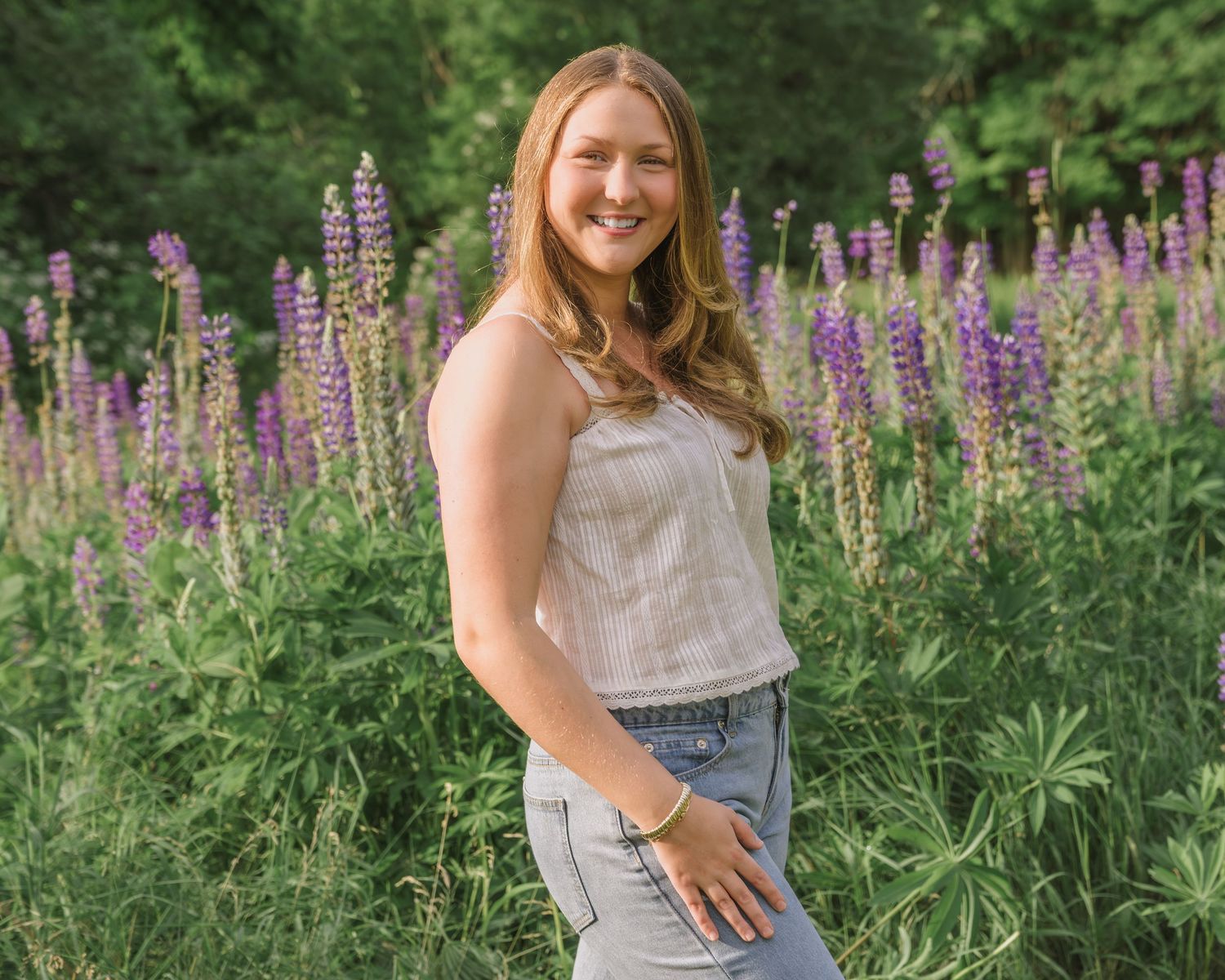 A person in a gray dress stands among vibrant purple lupine wildflowers in a lush green garden setting.