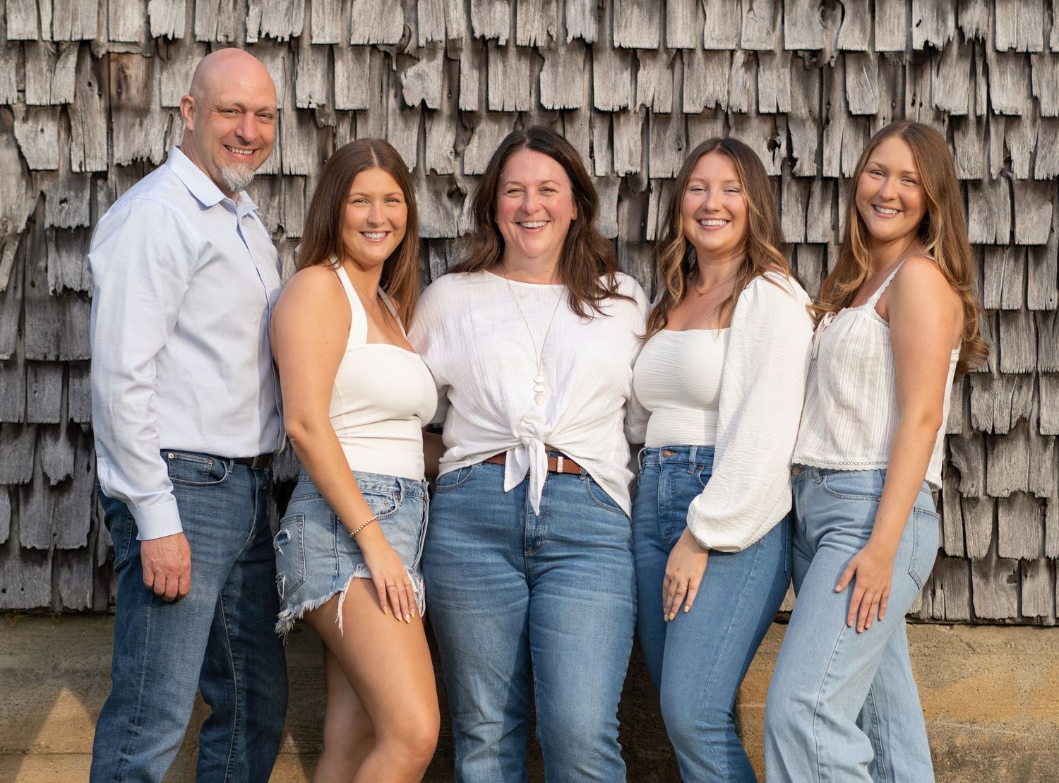 A group poses in coordinated blue jeans and white tops against a rustic wooden wall.