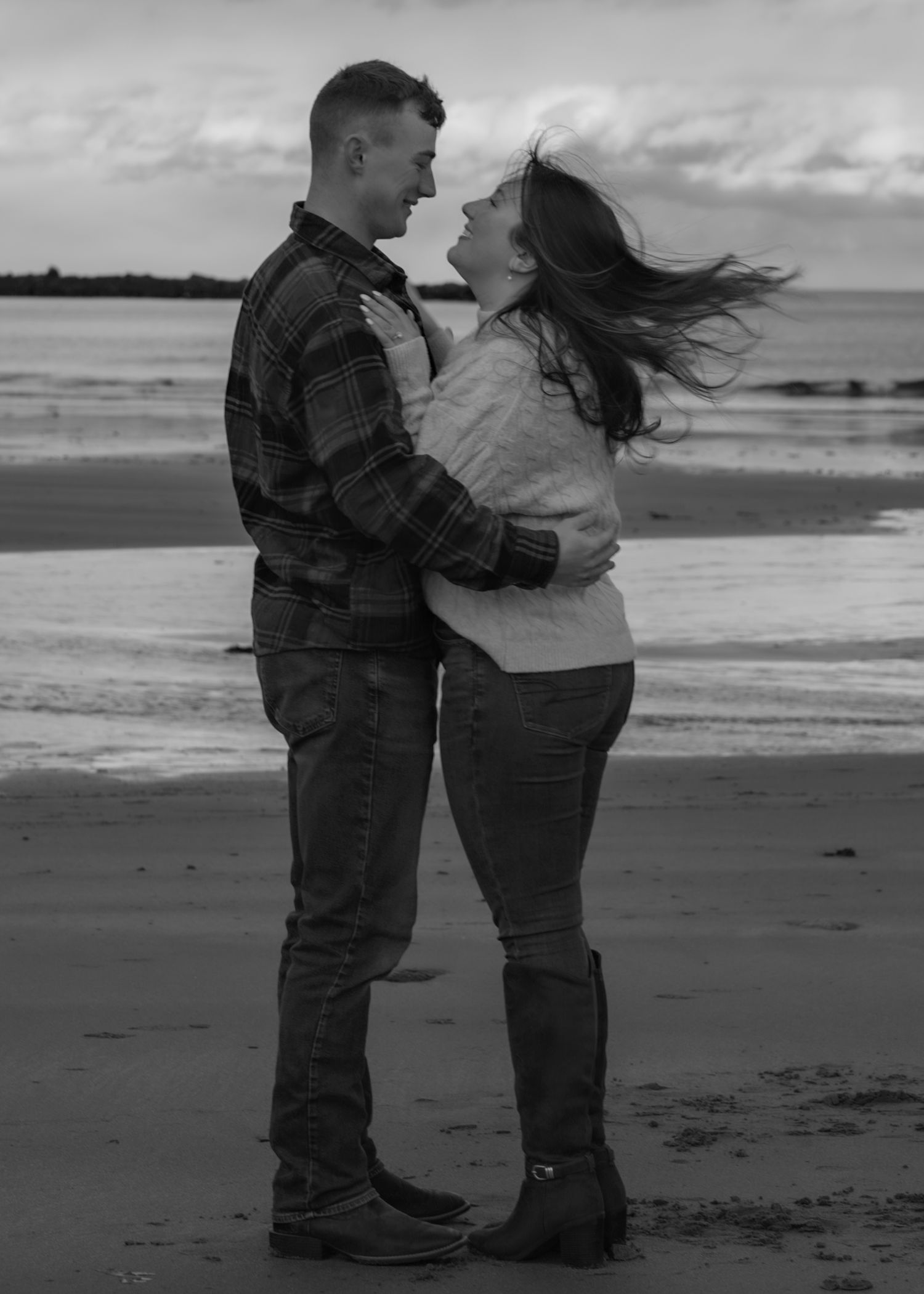 Couple embraces on windy beach in black and white photography.