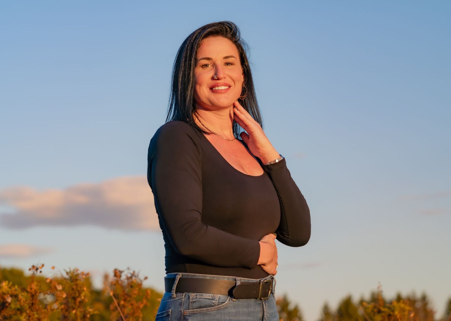 A person in a dark top poses outdoors during golden hour with autumn foliage in the background.