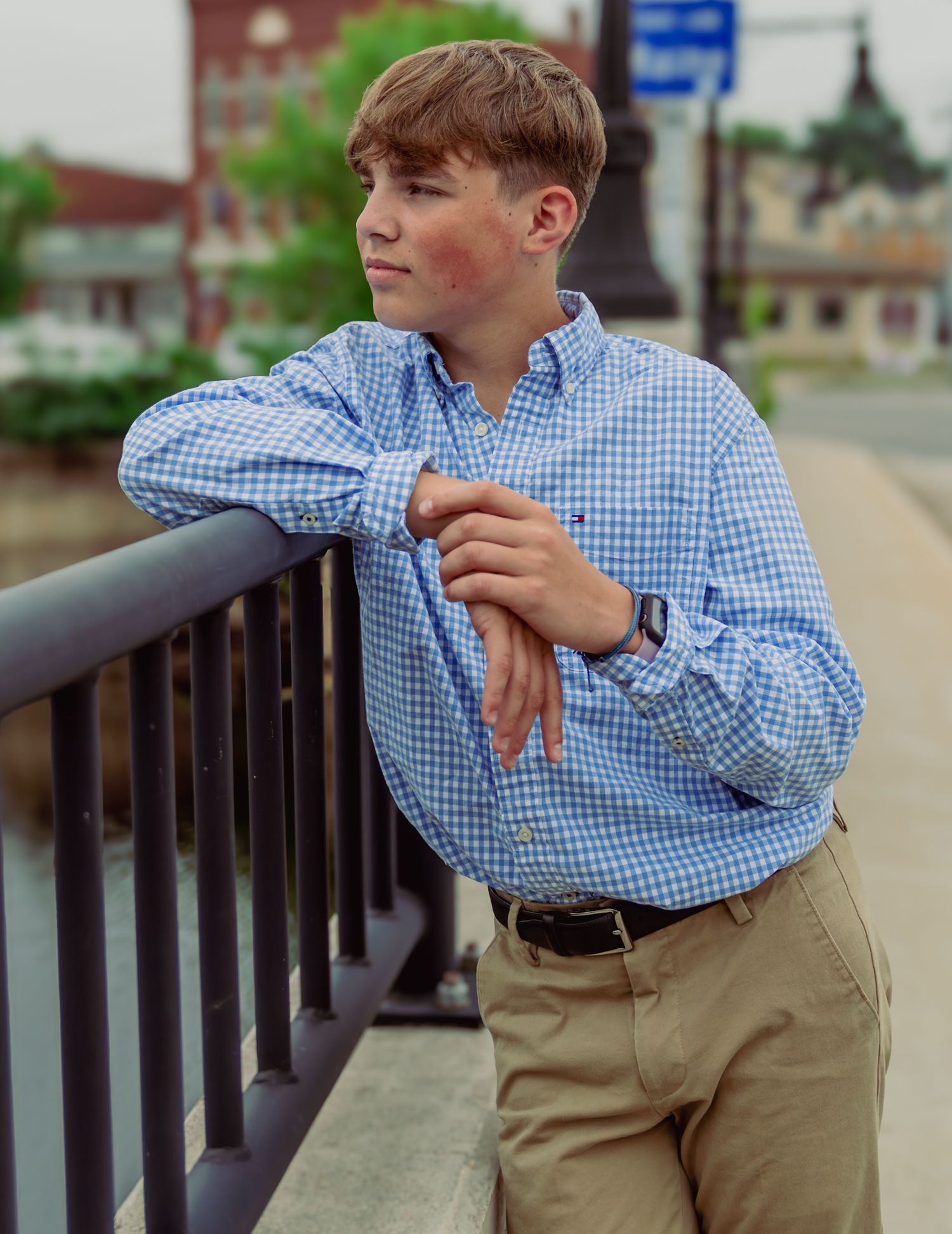 Young person in blue gingham shirt and khakis leaning against railing in casual outdoor portrait.