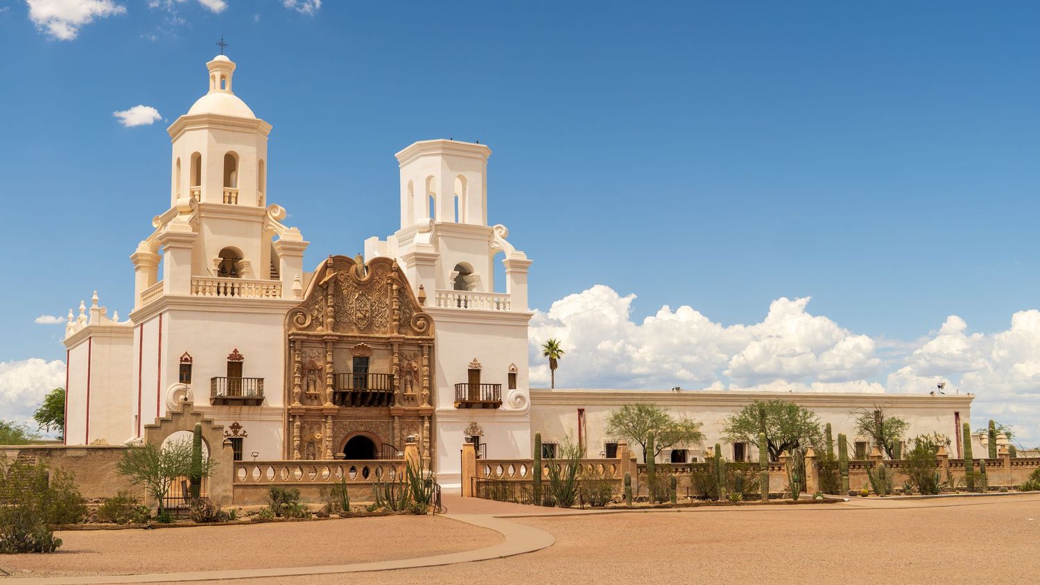 White mission church buildings with domes and towers stand against a bright blue desert sky.