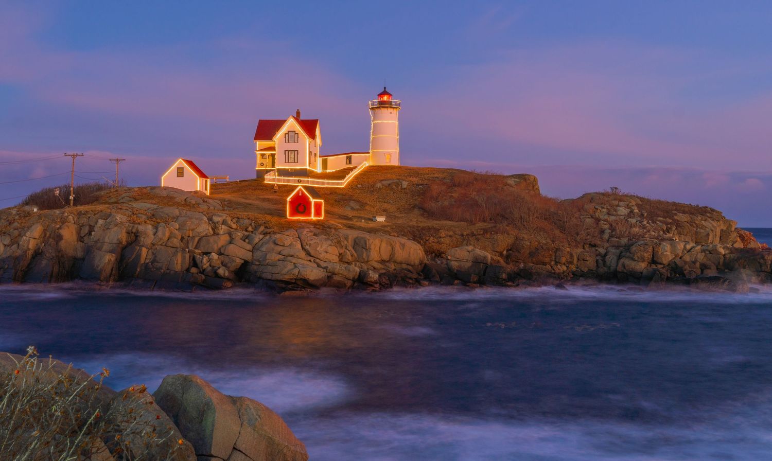 Nubble lighthouse and coastal buildings illuminated by dramatic sunset sky over rocky shoreline.