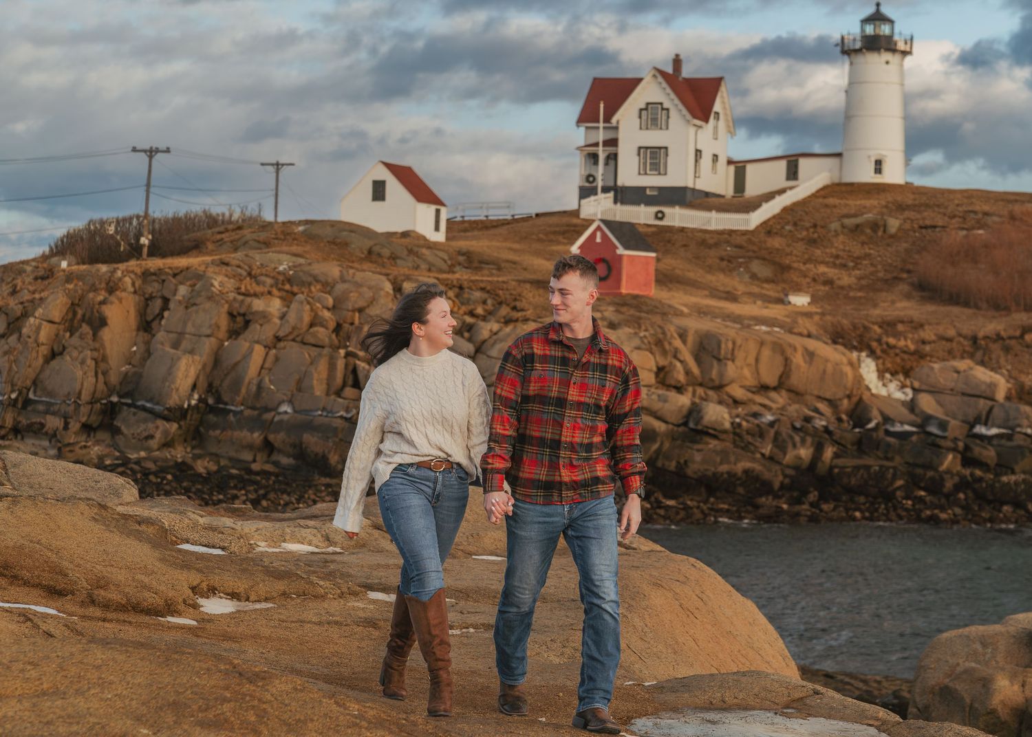 A couple walks along rocky coastal cliffs with a lighthouse in the background at sunset.