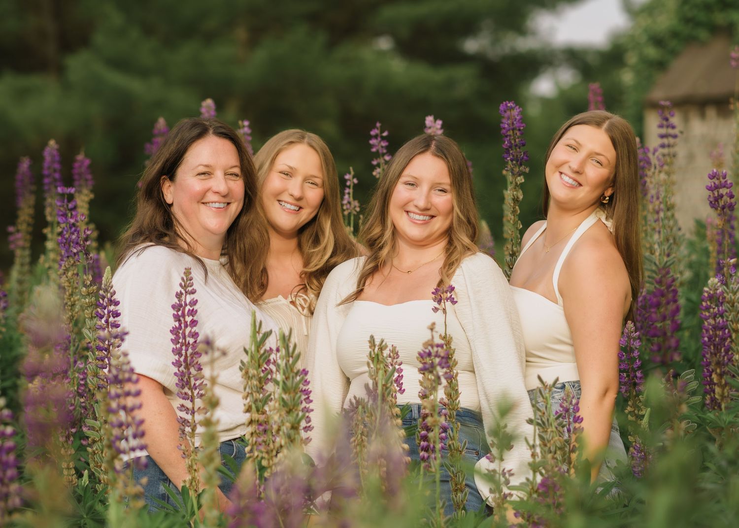 A group wearing white dresses poses joyfully among blooming purple lupine flowers in a garden setting.