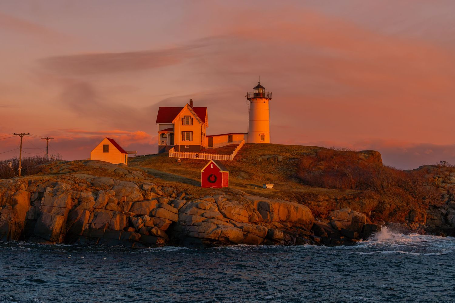 Nubble Lighthouse in Maine glows orange against rocky coast during dramatic sunset.