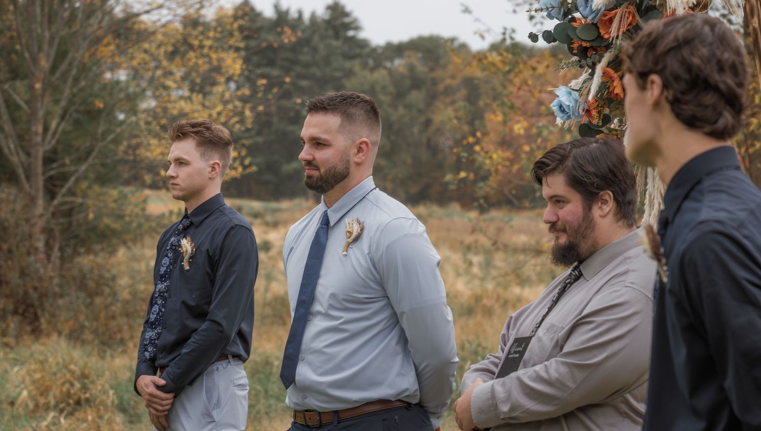 Group of groomsmen standing together in autumn field during wedding.