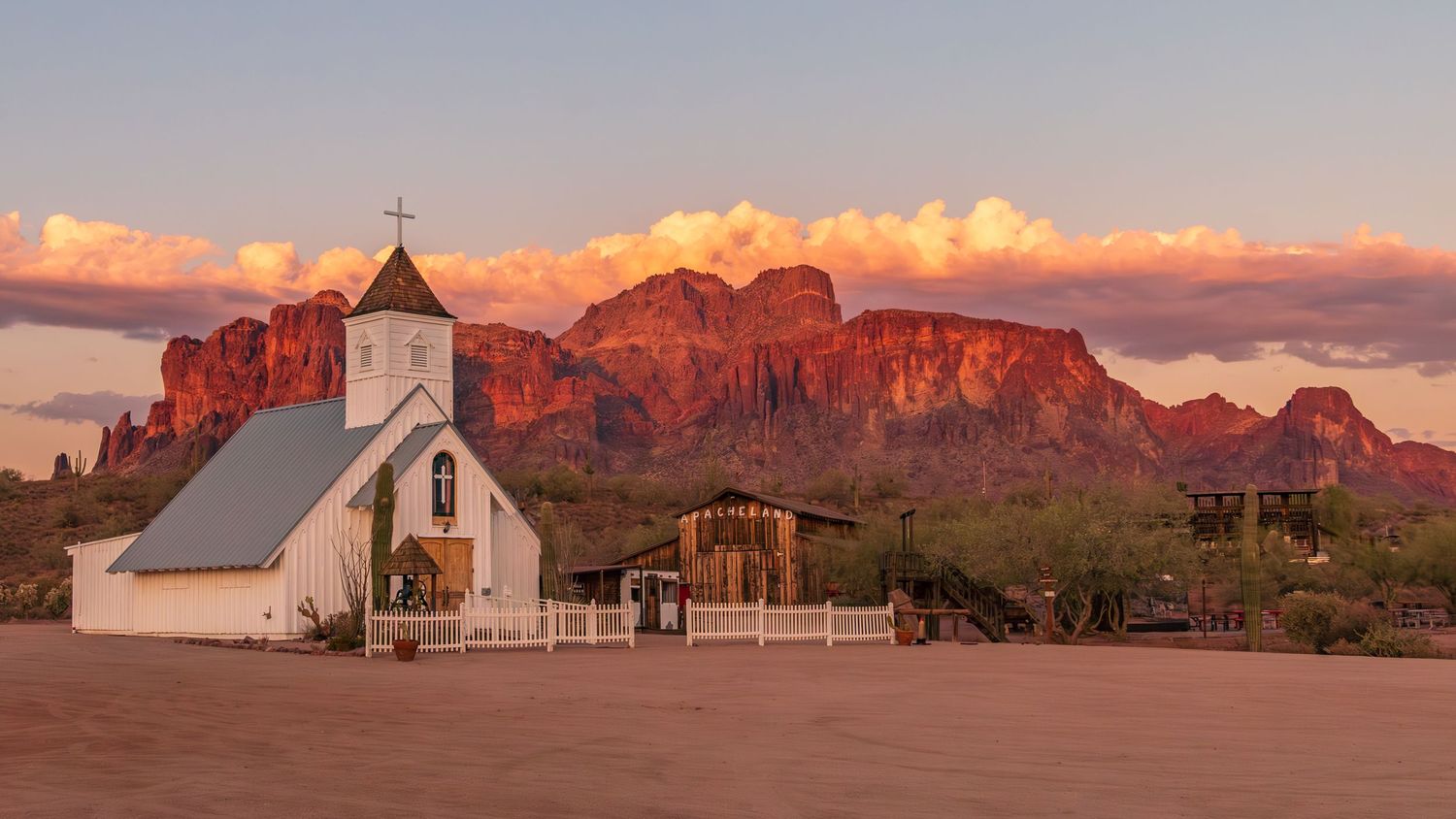 Desert mountains glow orange at sunset behind a white adobe mission church.