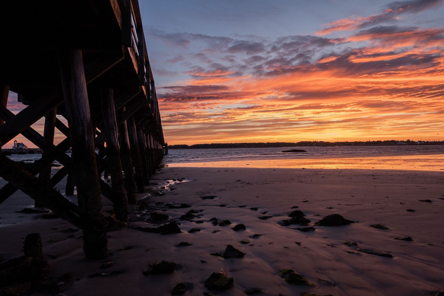 Dramatic sunset under a pier with silhouetted pillars and rocks on the beach.
