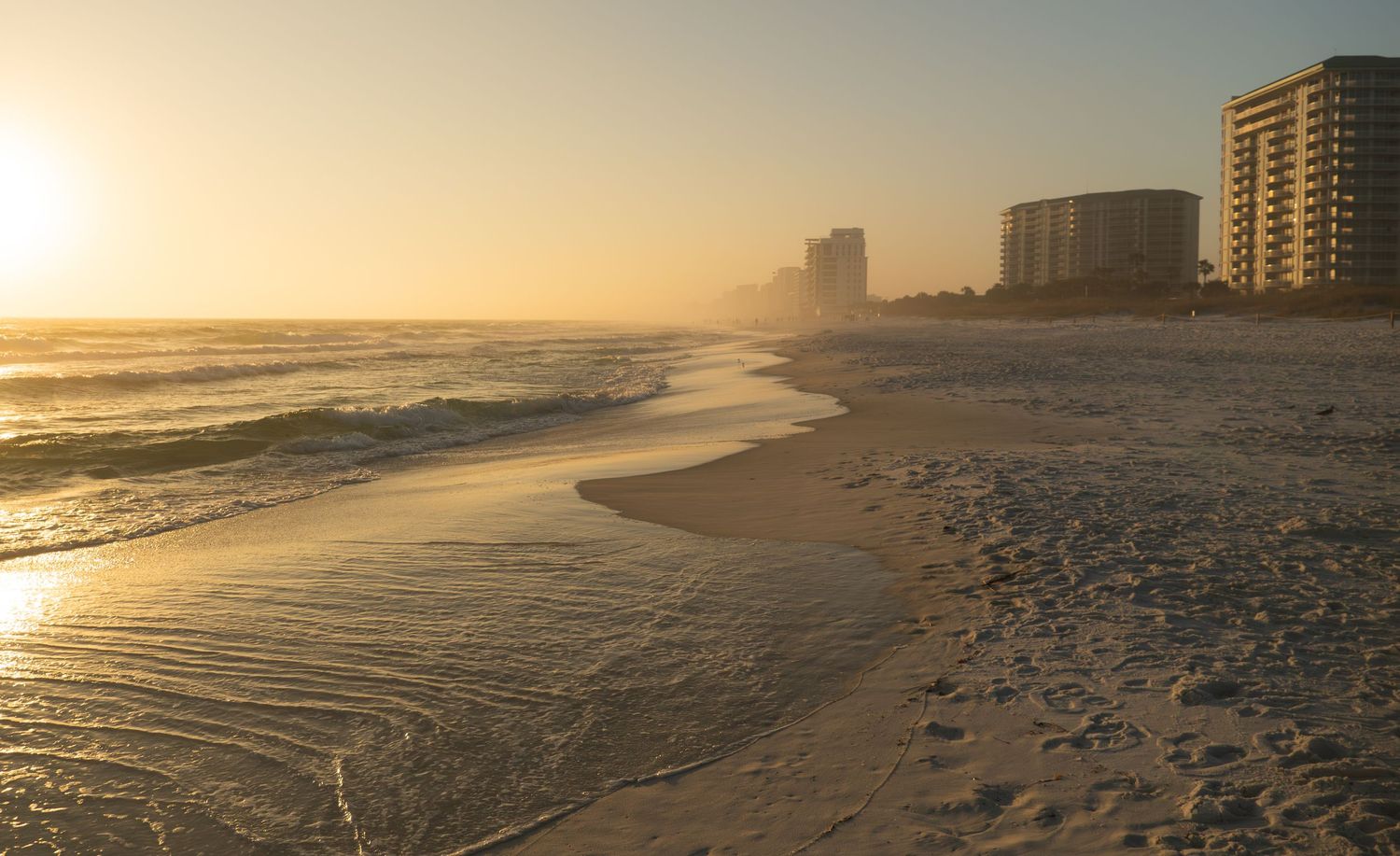 Golden sunset over a sandy beach with high-rise buildings visible through ocean mist.