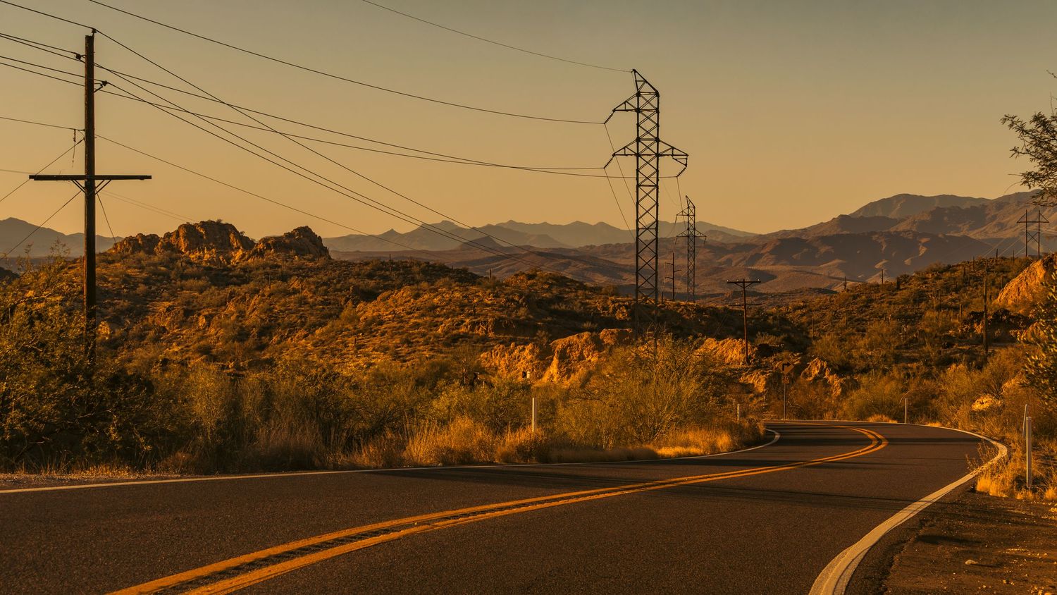Winding desert road with power lines and mountains silhouetted against a golden sunset.