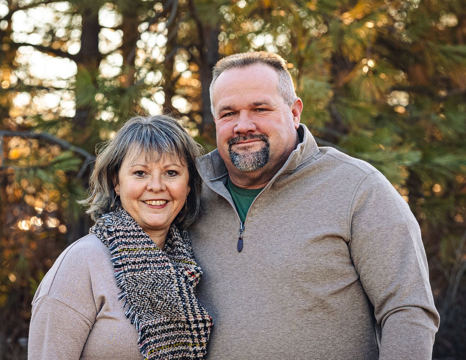 A couple in casual winter attire smiling and embracing outdoors during golden hour with trees in the background.