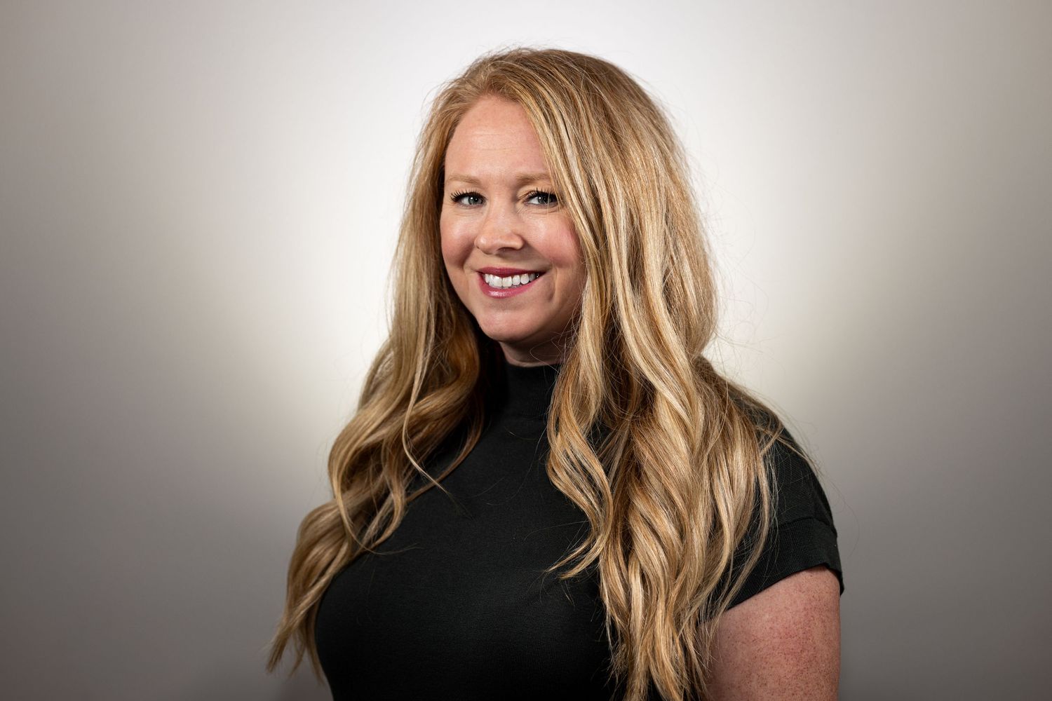 Professional headshot of someone with long blonde hair wearing a black turtleneck against a neutral background.