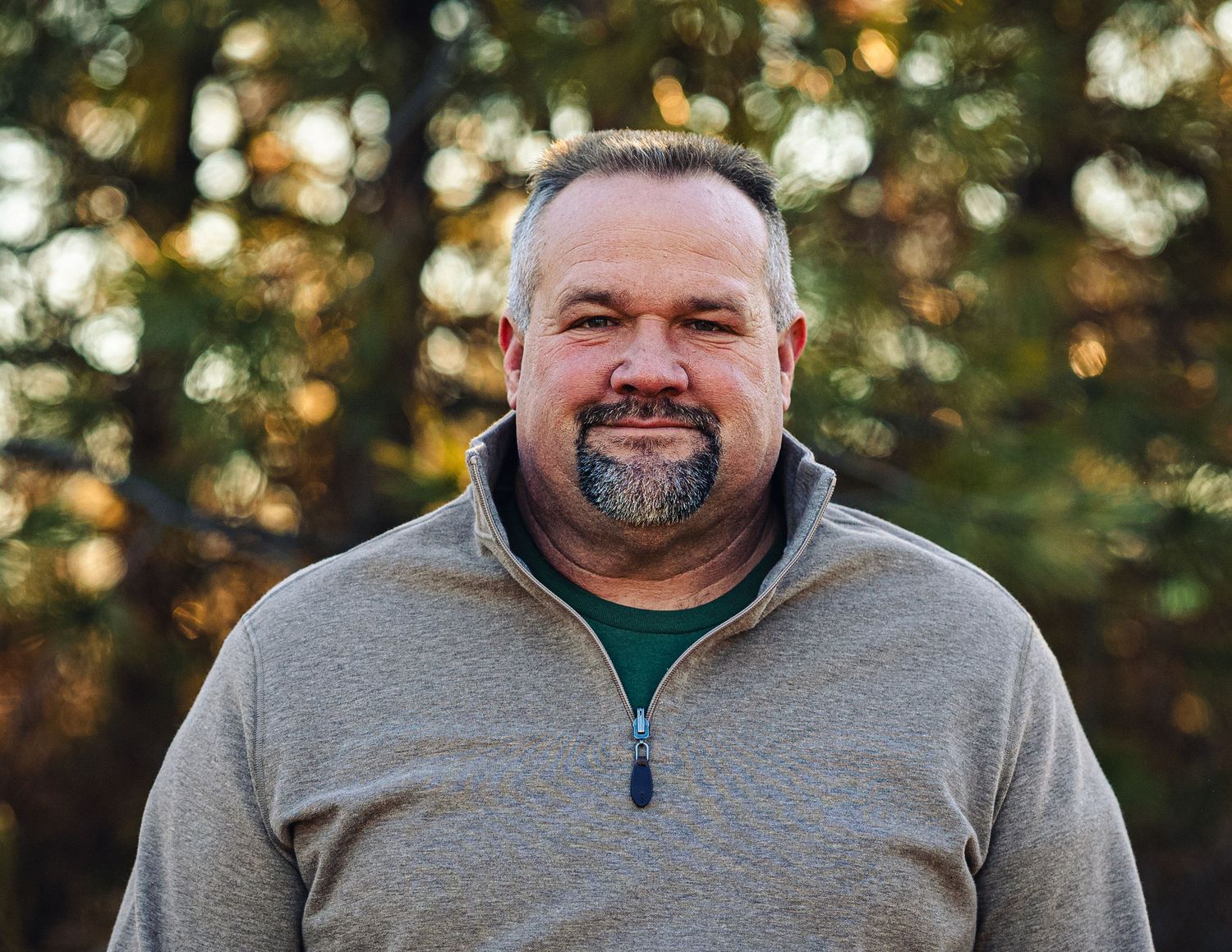 A person wearing a gray quarter-zip sweater smiles warmly while standing outdoors with blurred autumn foliage in background.