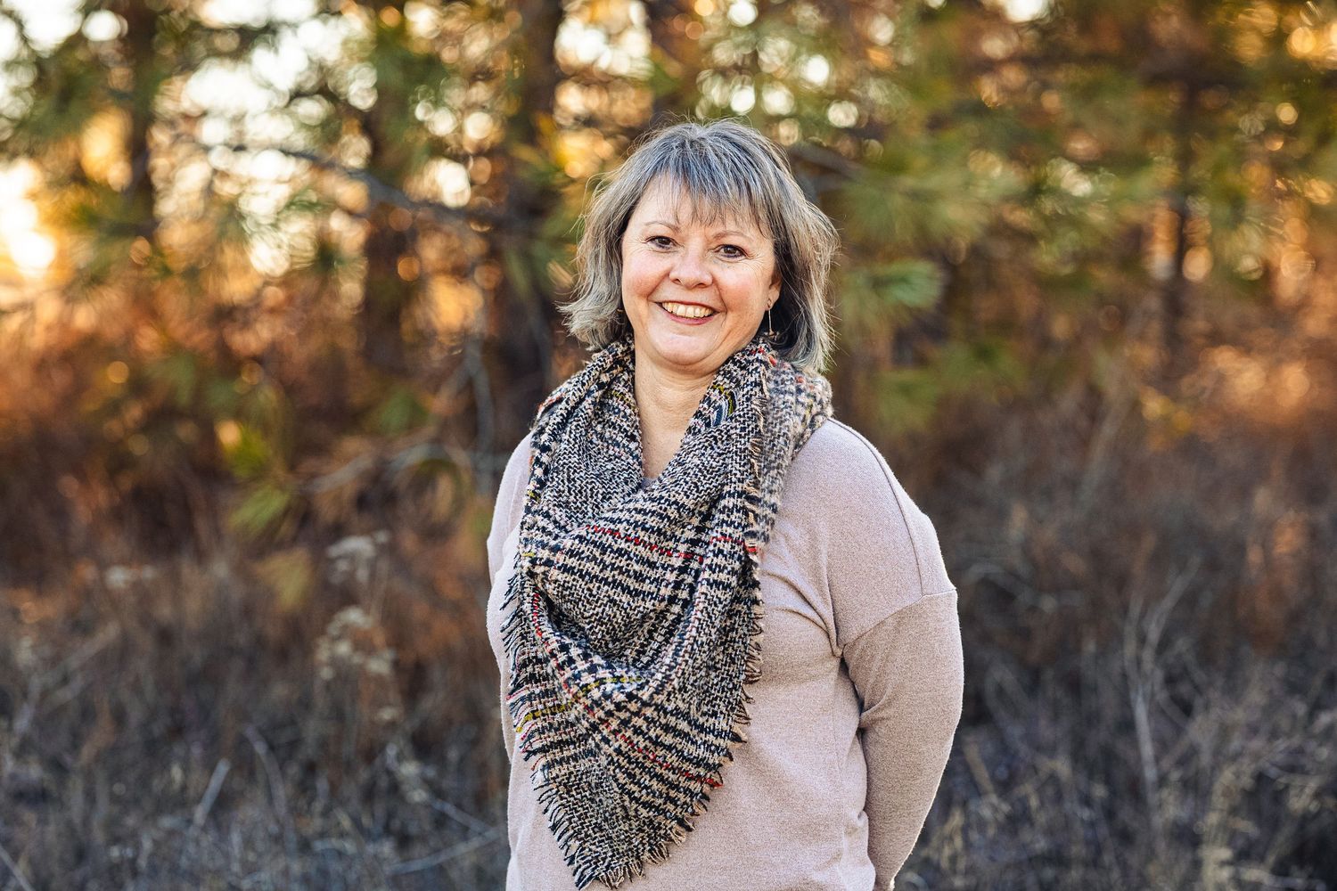 Woman in beige sweater and chunky knit infinity scarf poses outdoors during golden hour sunset.