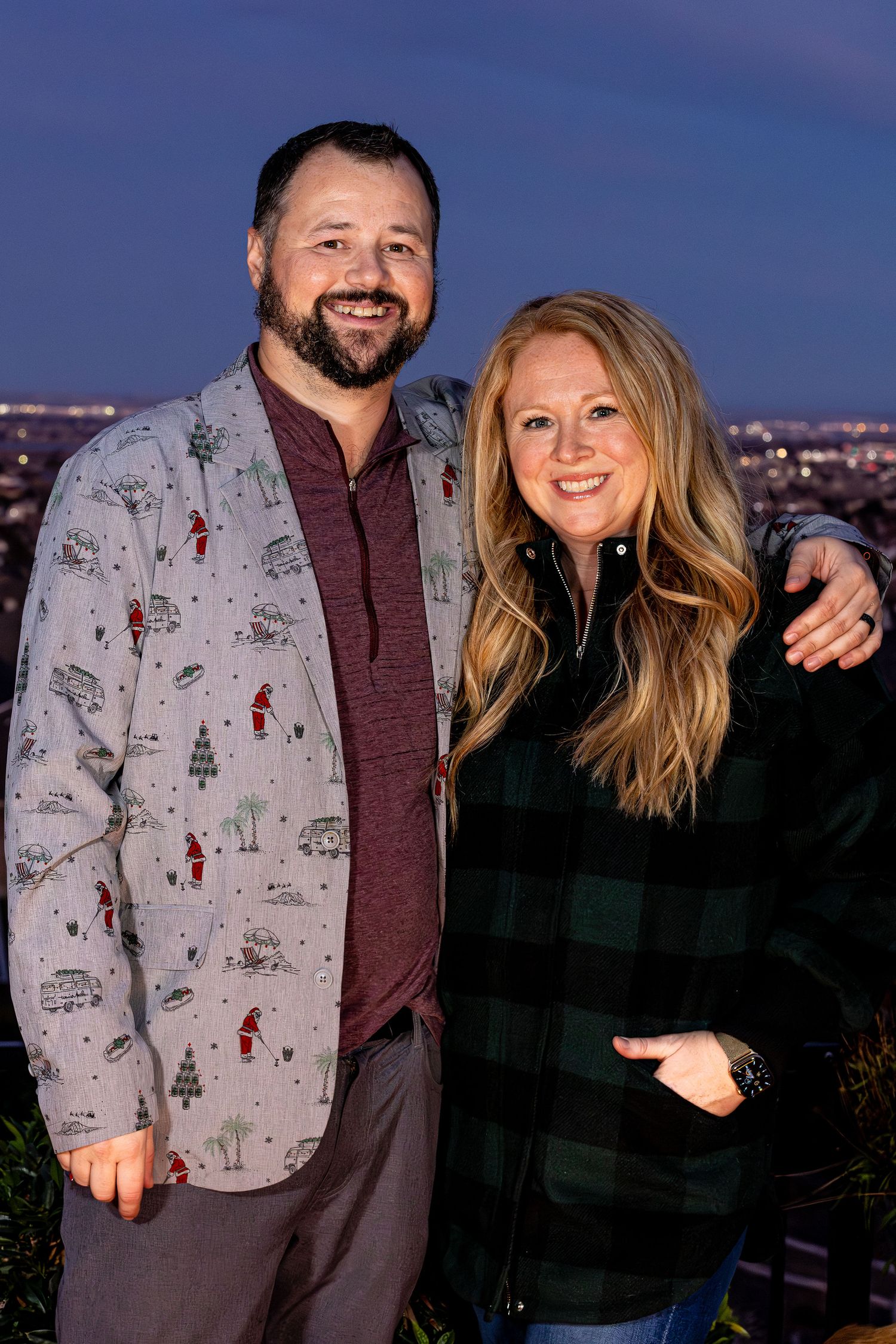 A couple poses together at night with city lights in the background, one wearing a patterned gray jacket.