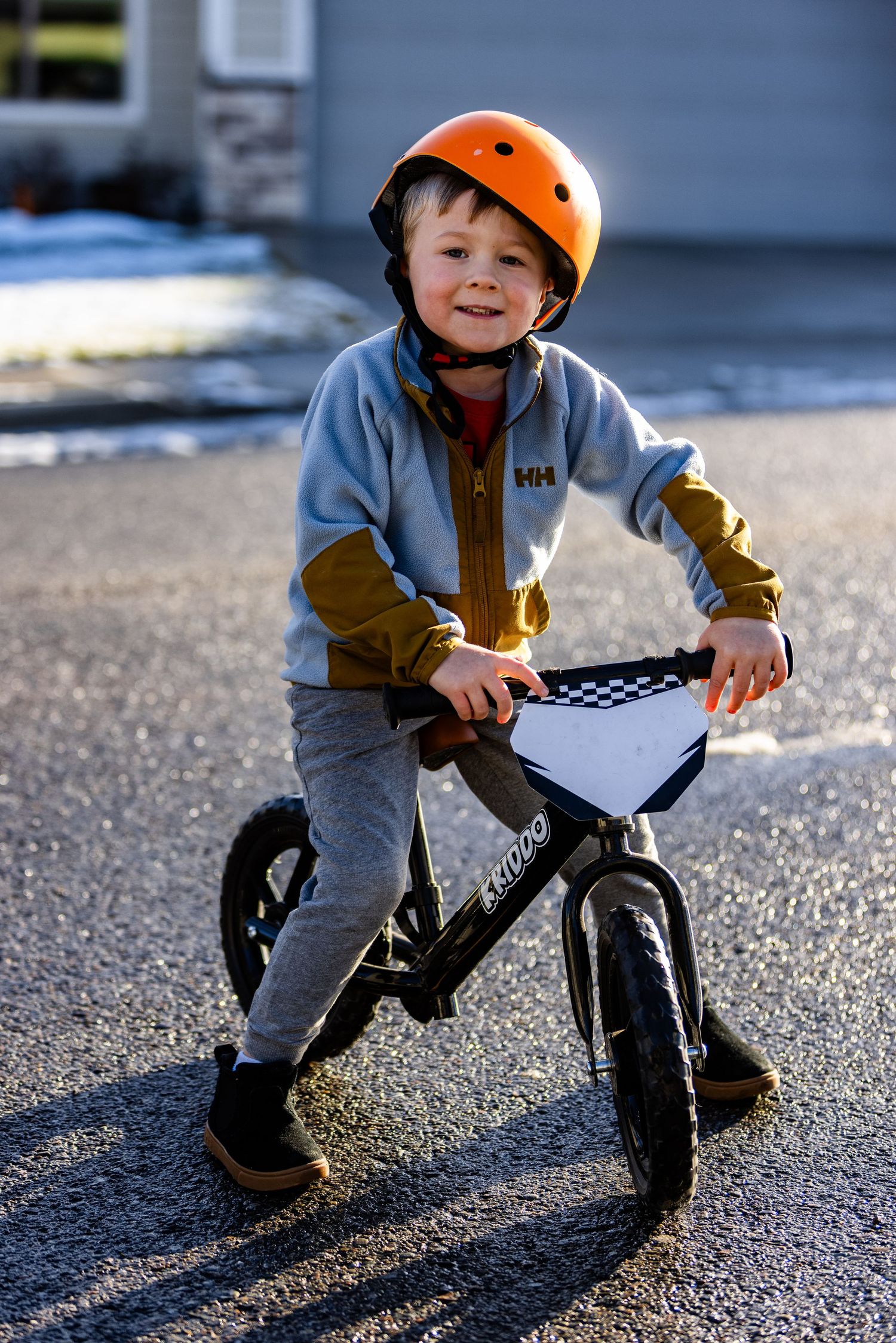 A small child rides a balance bike on pavement while wearing a bright orange helmet and gray clothing.