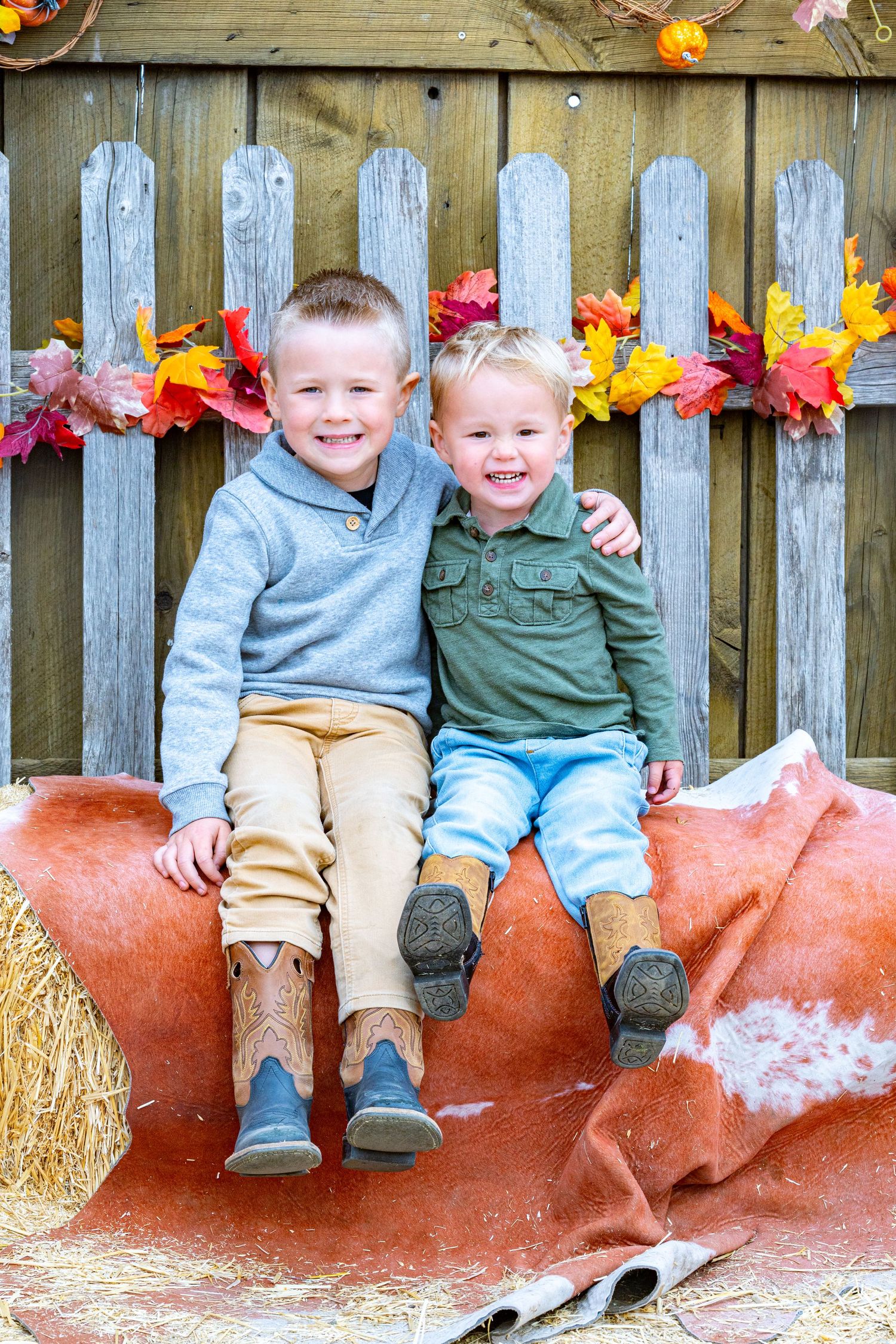 Two smiling toddler boys in cowboy boots sit together on a cowhide and hay bale in front of a rustic wooden fence with autumn leaf garland.