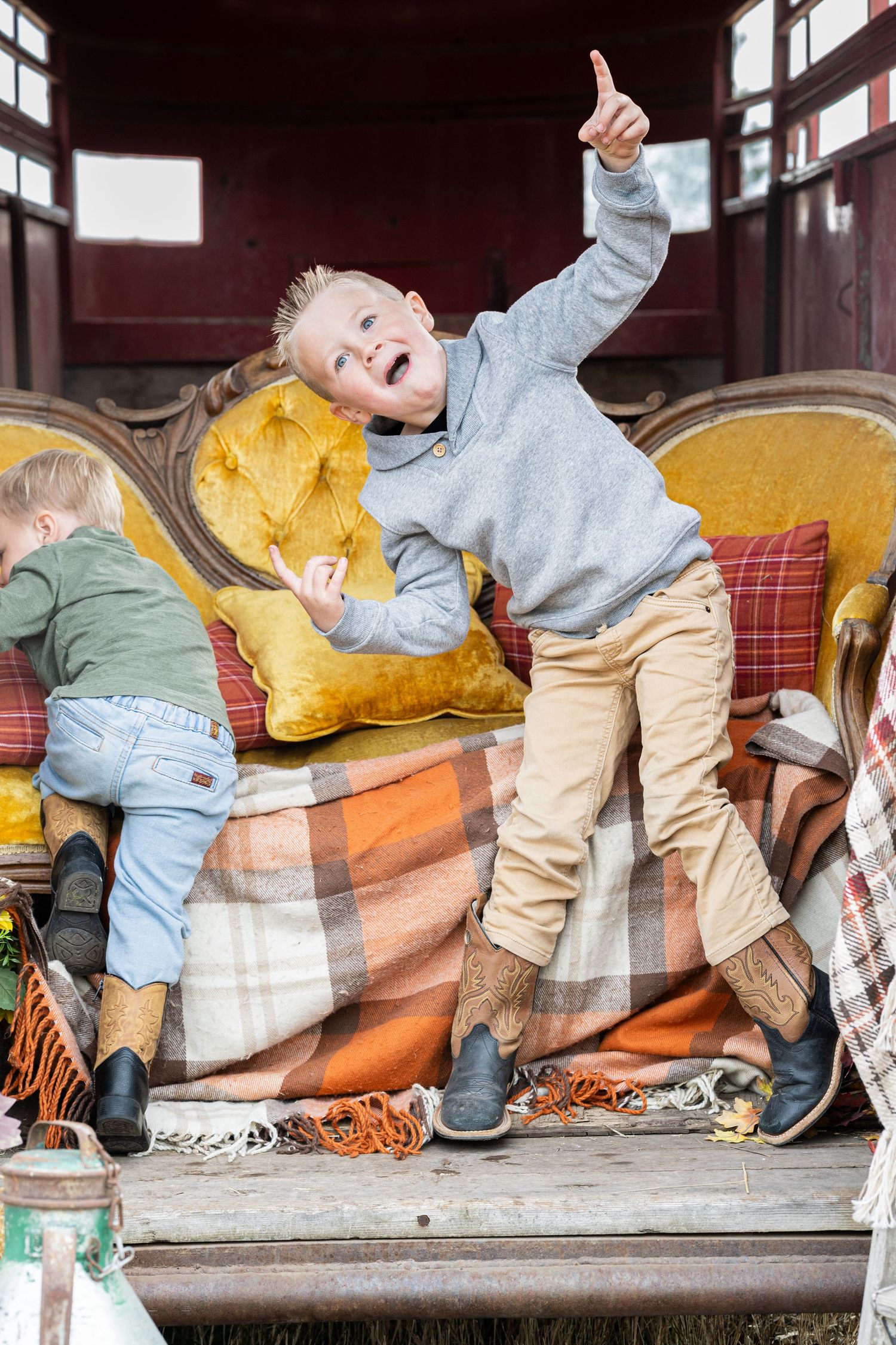 Playful blond boy in gray sweater and cowboy boots points upward while posing on a vintage yellow velvet sofa with plaid blankets.