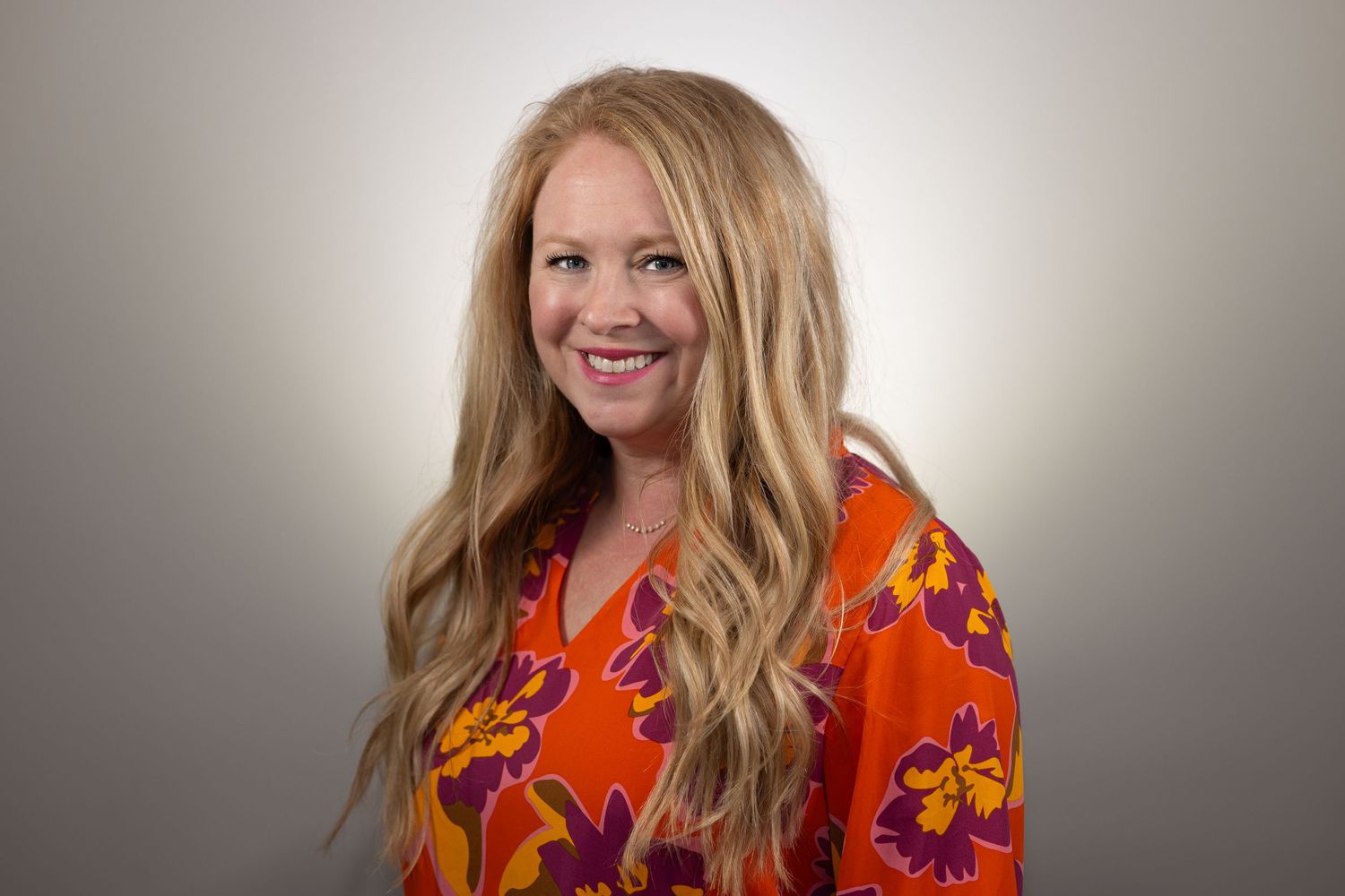 Professional headshot of someone wearing a vibrant orange floral blouse against a neutral background.