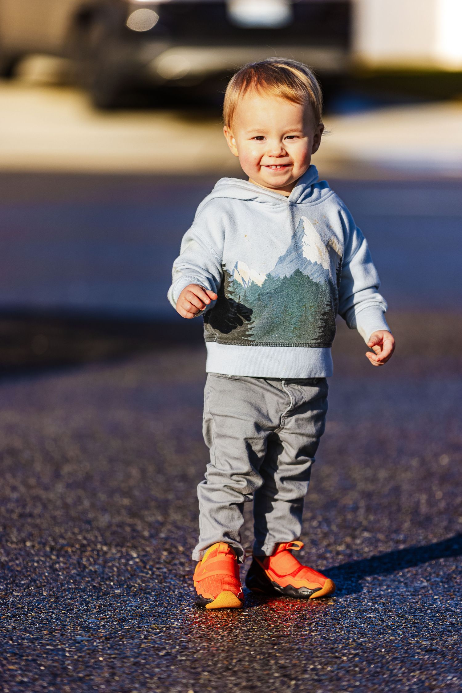 A small child wearing a light blue sweater and gray pants takes steps on pavement while wearing bright orange shoes.