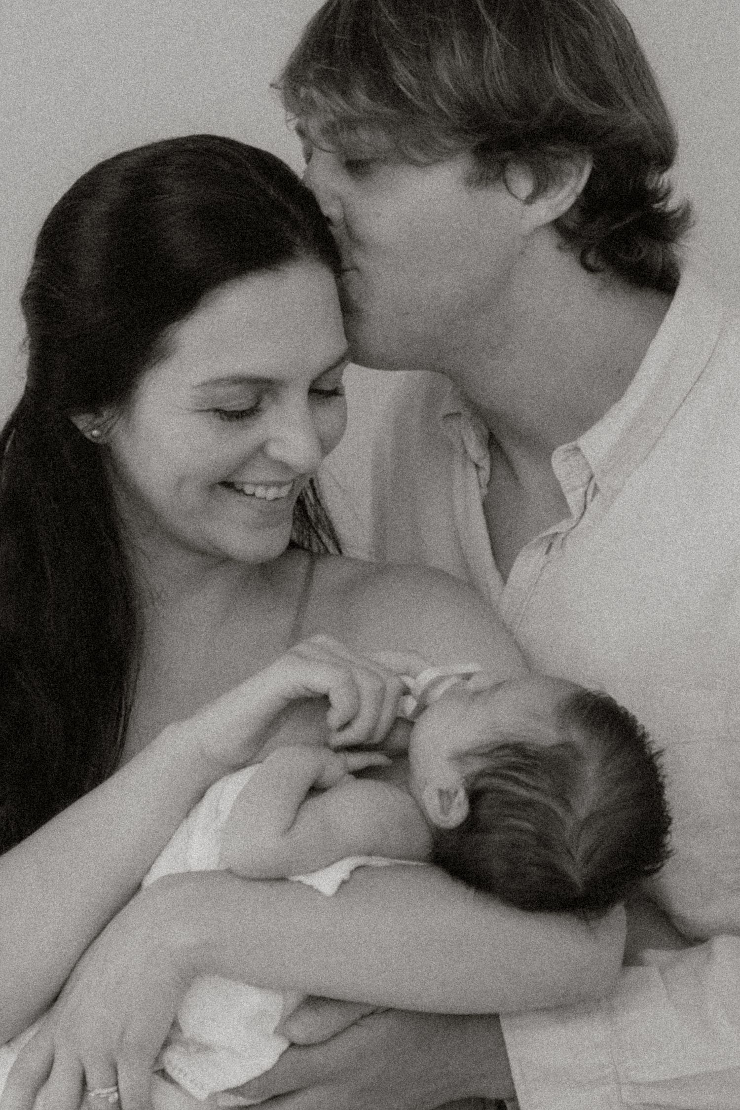 Black and white portrait of smiling new parents with father kissing mother's head, holding newborn.