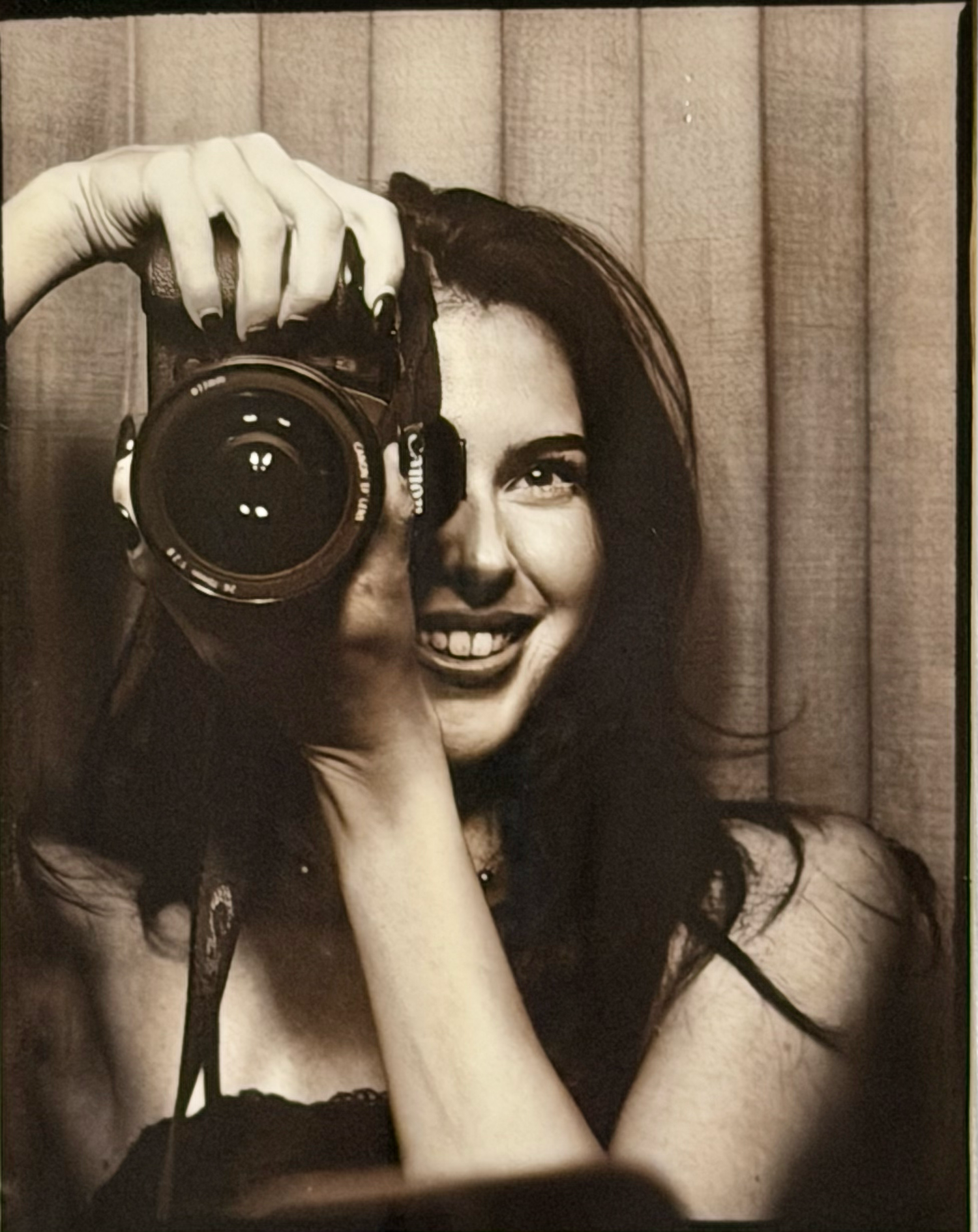 Sepia-toned photo booth portrait of a dark-haired woman smiling while holding a DSLR camera.