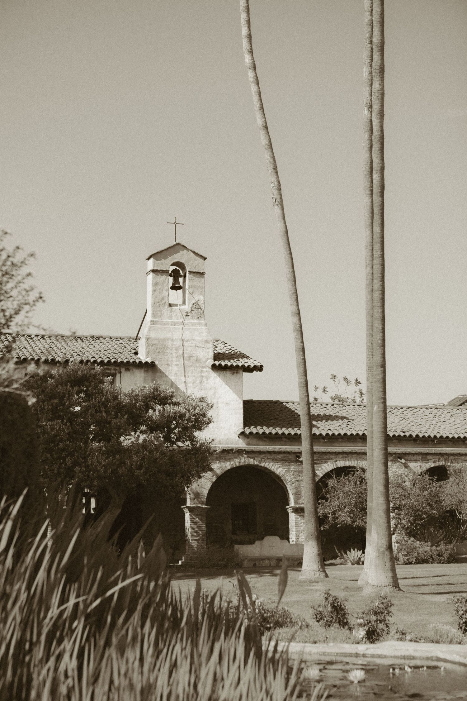 Sepia-toned photo of historic Spanish mission bell tower with palm trees and arched entrance.