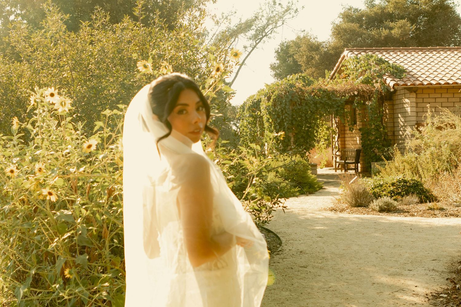 Bride in a white gown and veil posing in a sun-drenched garden path.