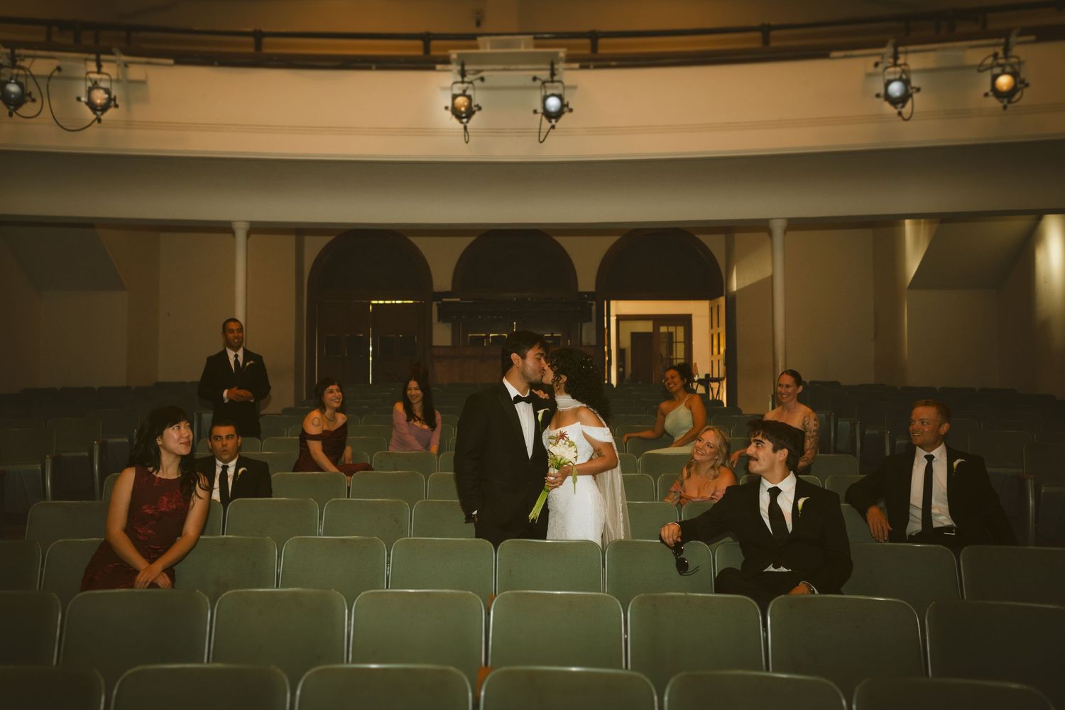 Bride and groom share a kiss in a vintage theater with wedding guests seated in green velvet chairs.