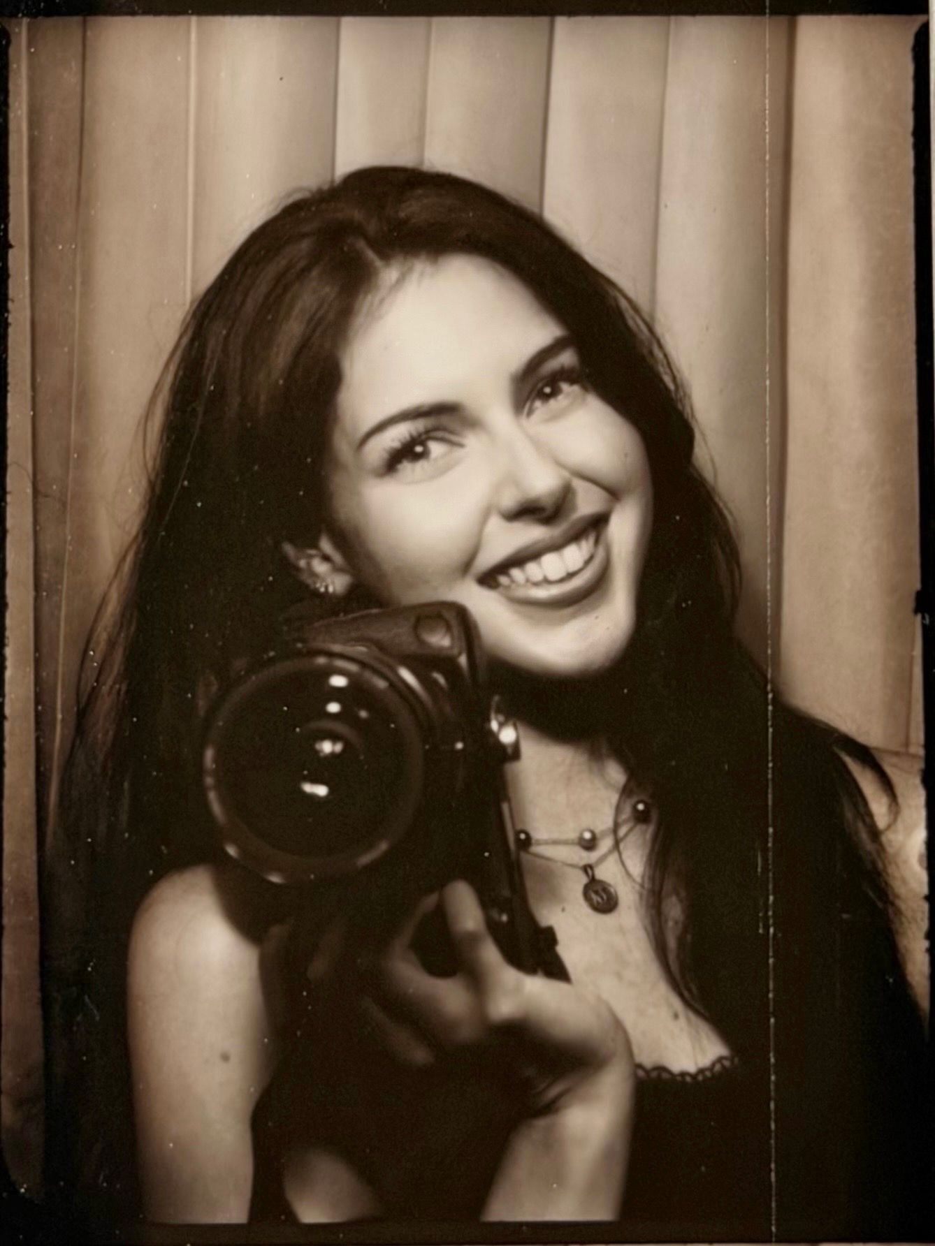 Vintage sepia photo booth portrait of a smiling brunette woman holding a DSLR camera toward viewer.
