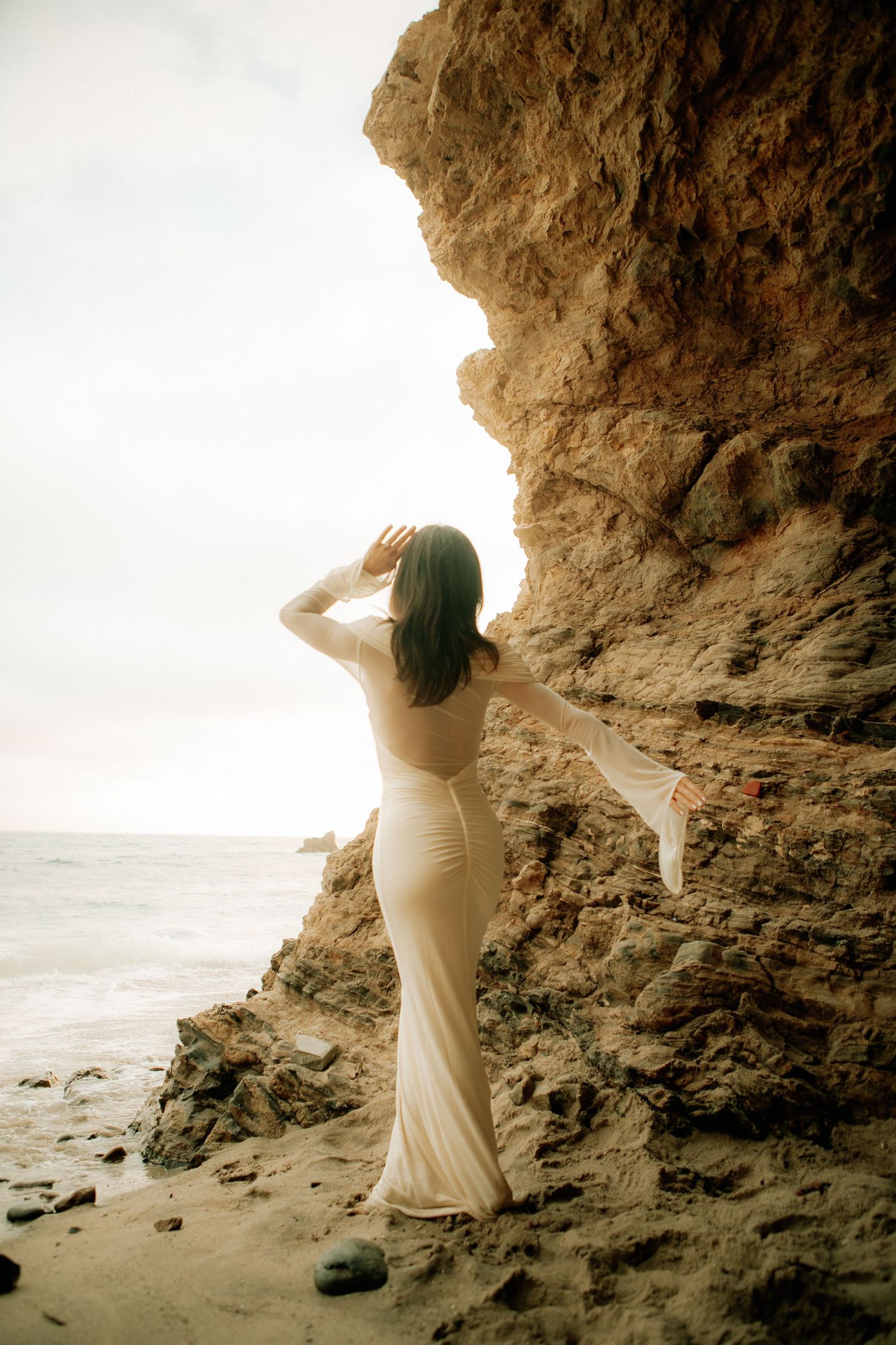 Woman in white fitted gown standing against rocky coastal cliffs gazing toward ocean.